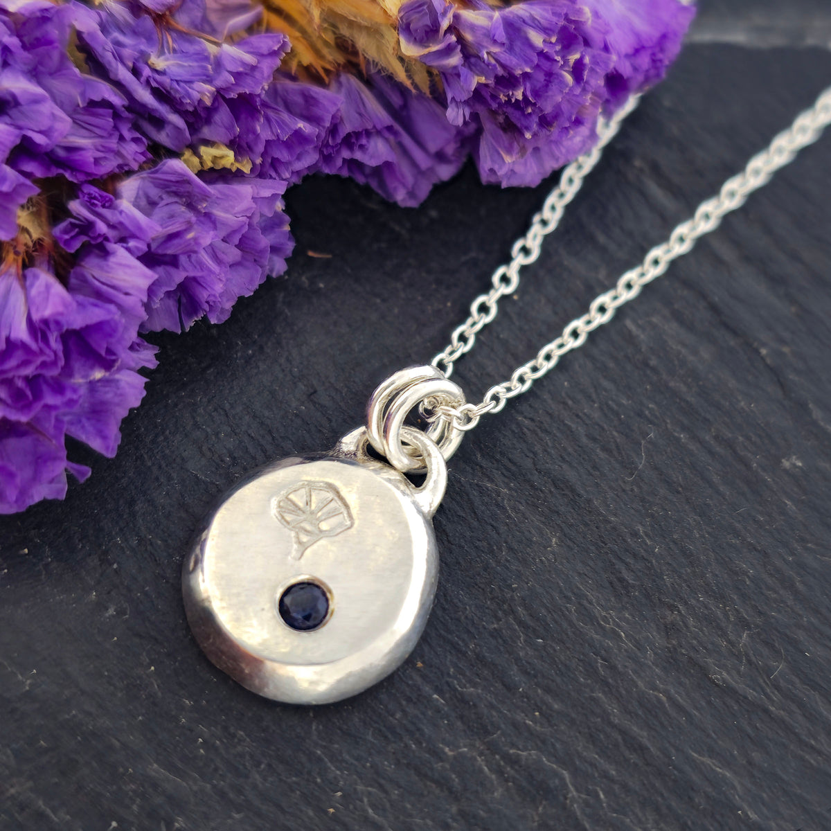A handmade silver pebble pendant necklace featuring a disc-shaped charm with a small dark blue sapphire gemstone and an engraved morning glory flower design, suspended from a delicate silver chain against a black slate background with purple flowers. Mijoux Jewellery.