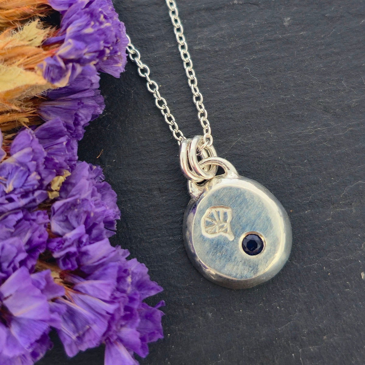 A handmade silver pebble pendant necklace featuring a disc-shaped charm with a small dark blue sapphire gemstone and an engraved morning glory flower design, suspended from a delicate silver chain against a black slate background with purple flowers. Mijoux Jewellery.