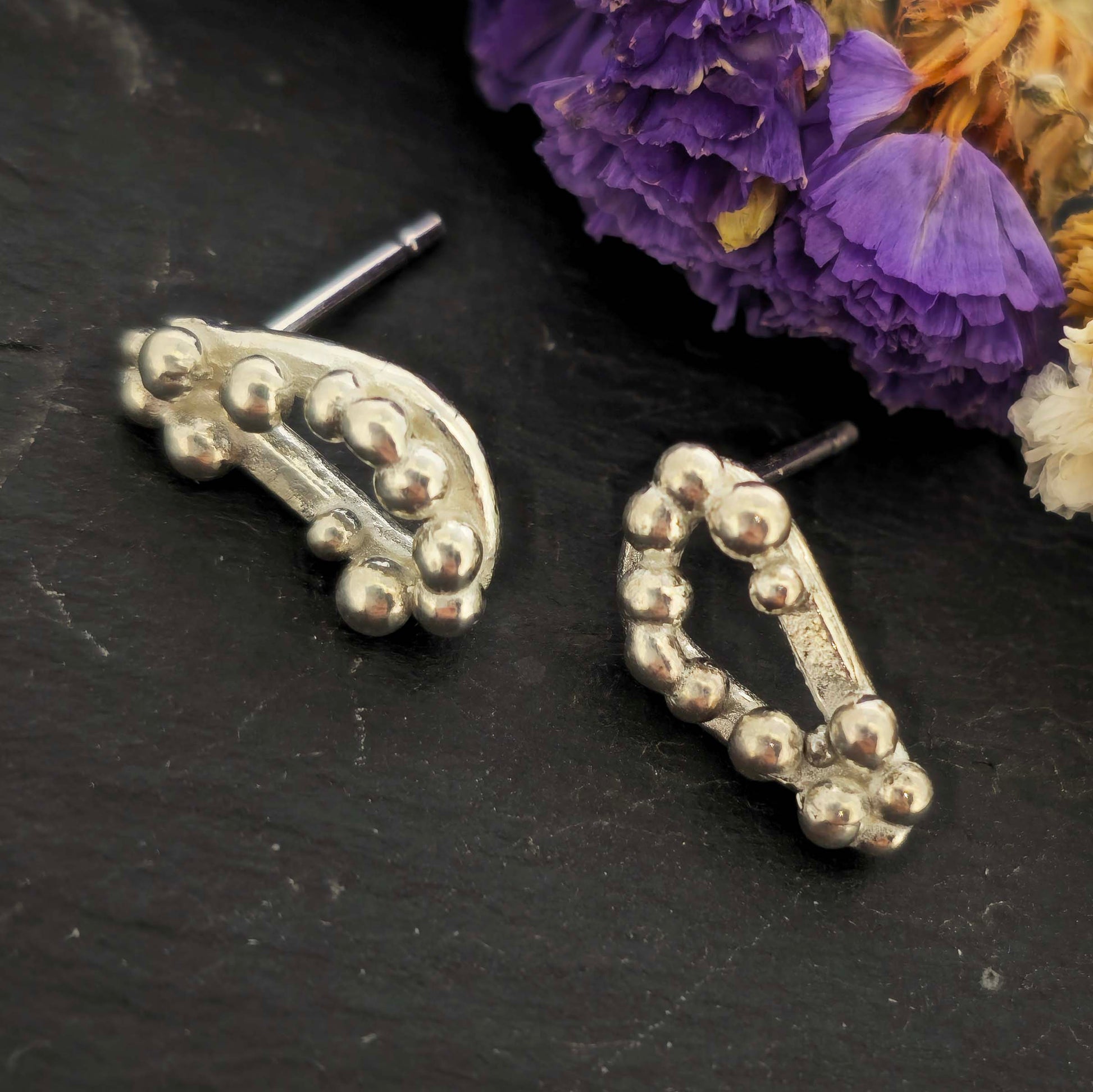 Silver asymmetric stud earrings with an open soft triangle shape and silver balls on a black background with dried flowers.