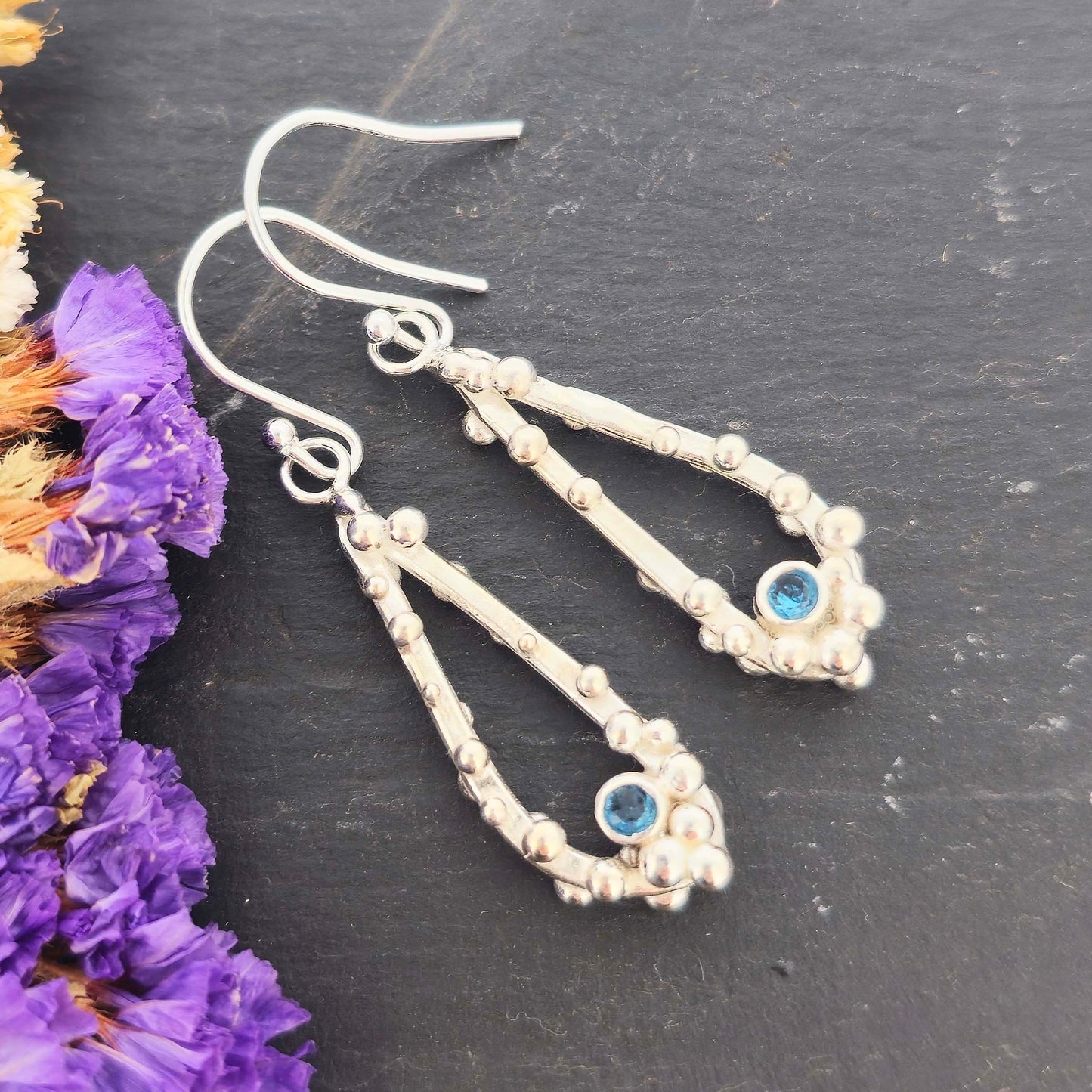 Silver drop earrings with open asymmetrical teardrop shape and silver balls and round blue gemstones. On a black background with dried flowers.
