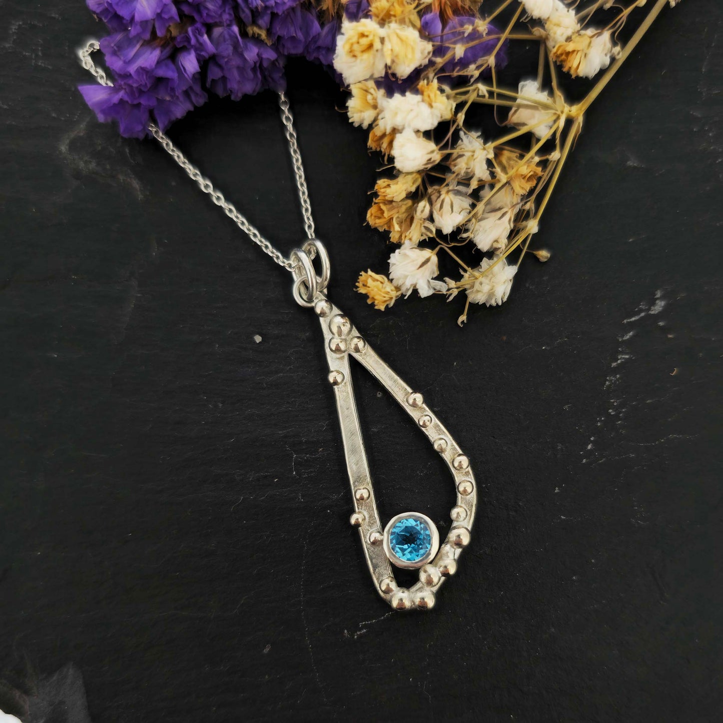Silver asymmetrical soft open triangle pendant with silver balls and a blue topaz gemstone on a silver chain. Shown on a black background with dried flowers.