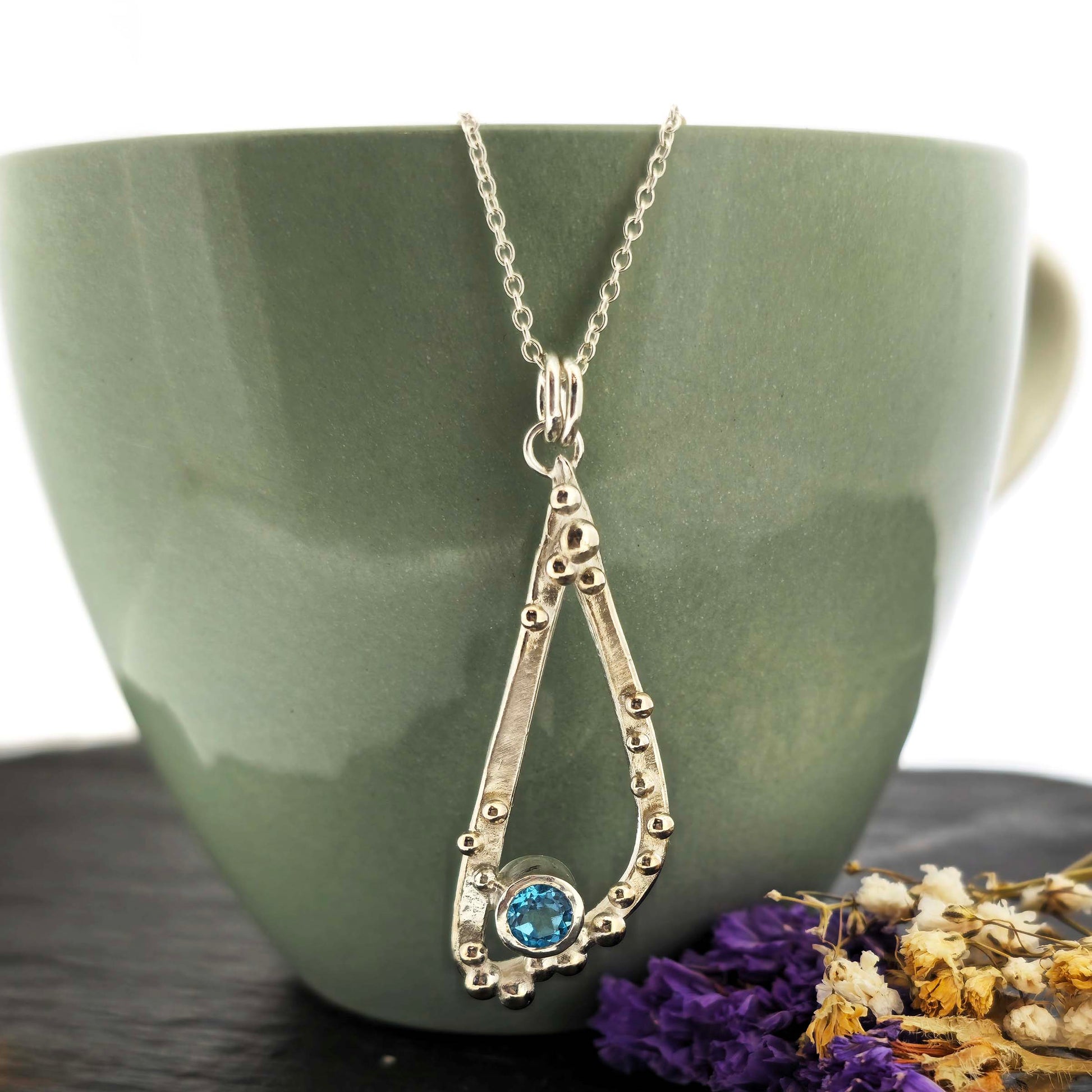 Silver asymmetrical soft open triangle pendant with silver balls and a blue topaz gemstone on a silver chain. Shown on a green cup with dried flowers.