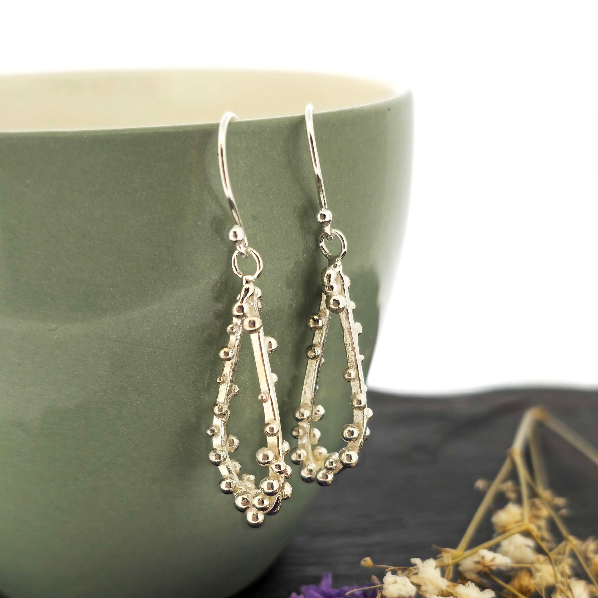 Silver drop earrings in a asymmetrical open teardrop shape with silver balls. Hung on a green bowl with dried flowers.