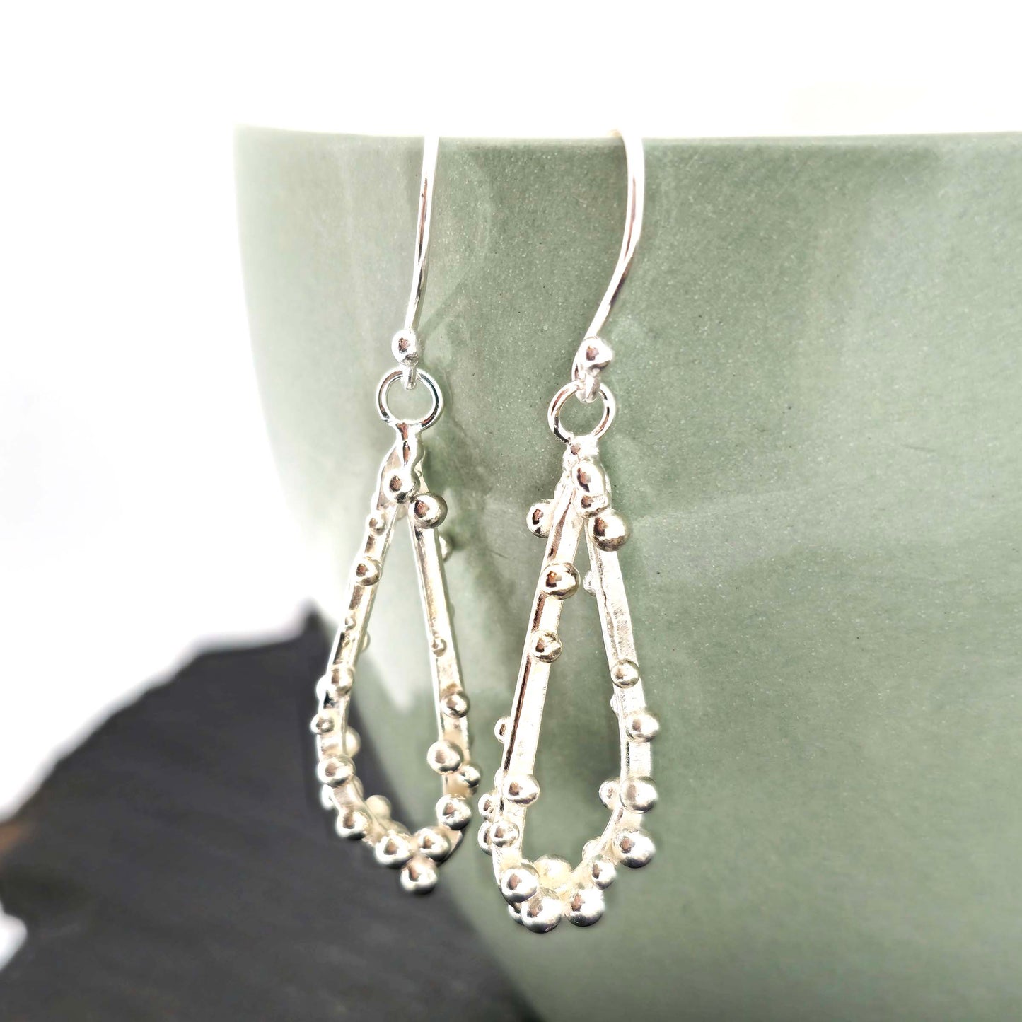 Silver drop earrings in a asymmetrical open teardrop shape with silver balls. Hung on a green bowl with dried flowers.