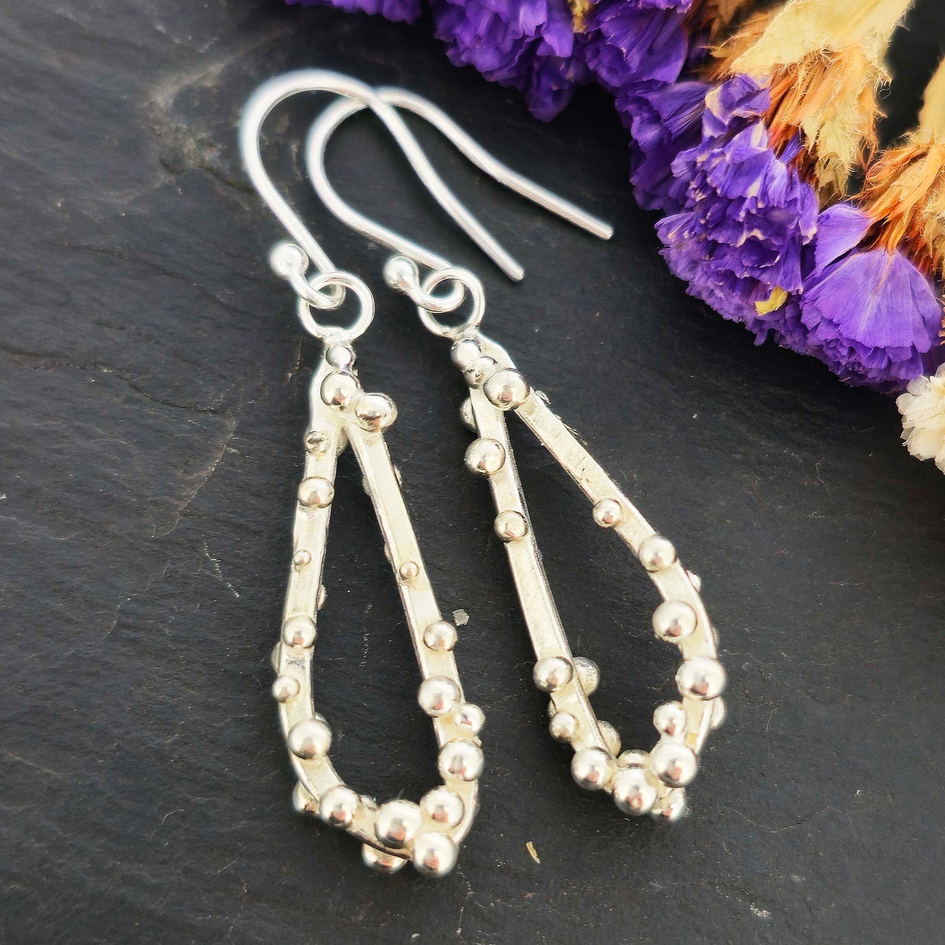 Silver drop earrings in a asymmetrical open teardrop shape with silver balls. Shown on a black background with dried flowers.