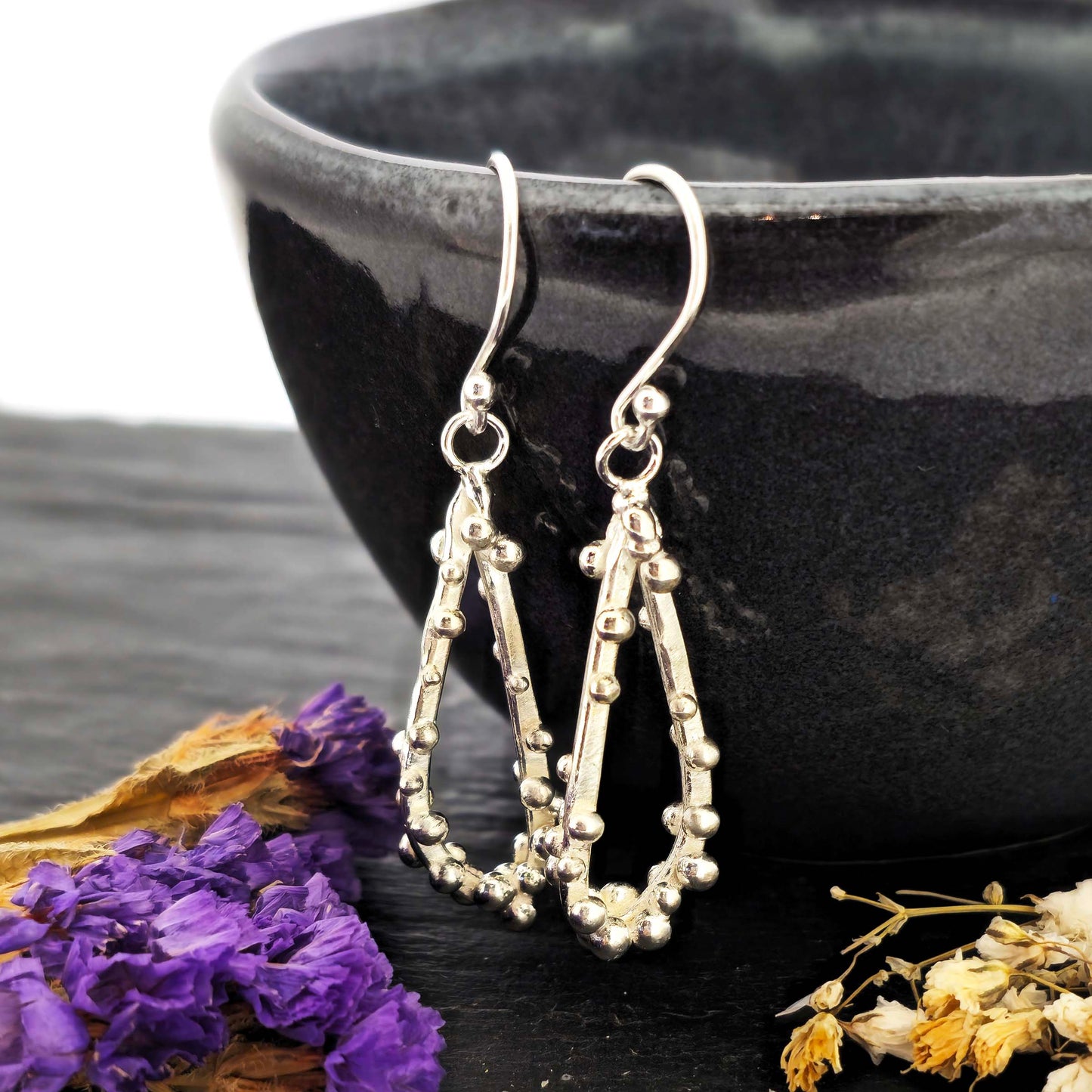 Silver drop earrings in a asymmetrical open teardrop shape with silver balls. Hung on a black bowl with dried flowers.