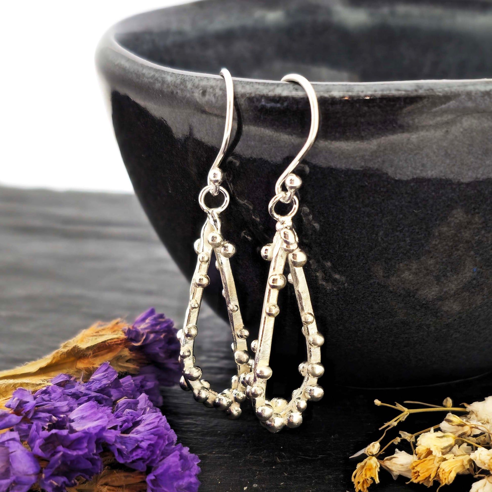 Silver drop earrings in a asymmetrical open teardrop shape with silver balls. Hung on a black bowl with dried flowers.
