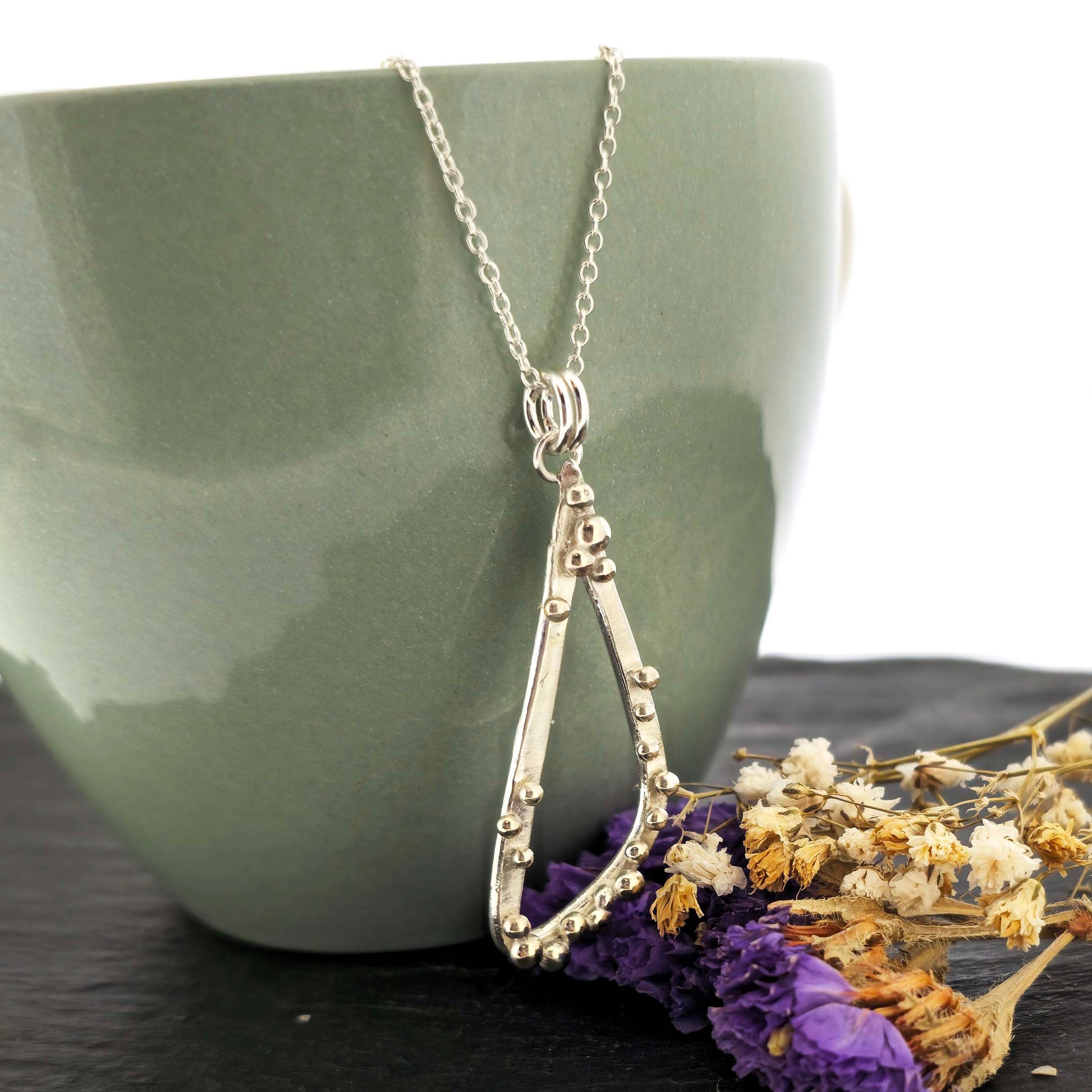 A silver asymmetrical pendant with a soft open triangle shape and silver balls, suspended from a silver chain. Shown on a green bowl with dried flowers. 