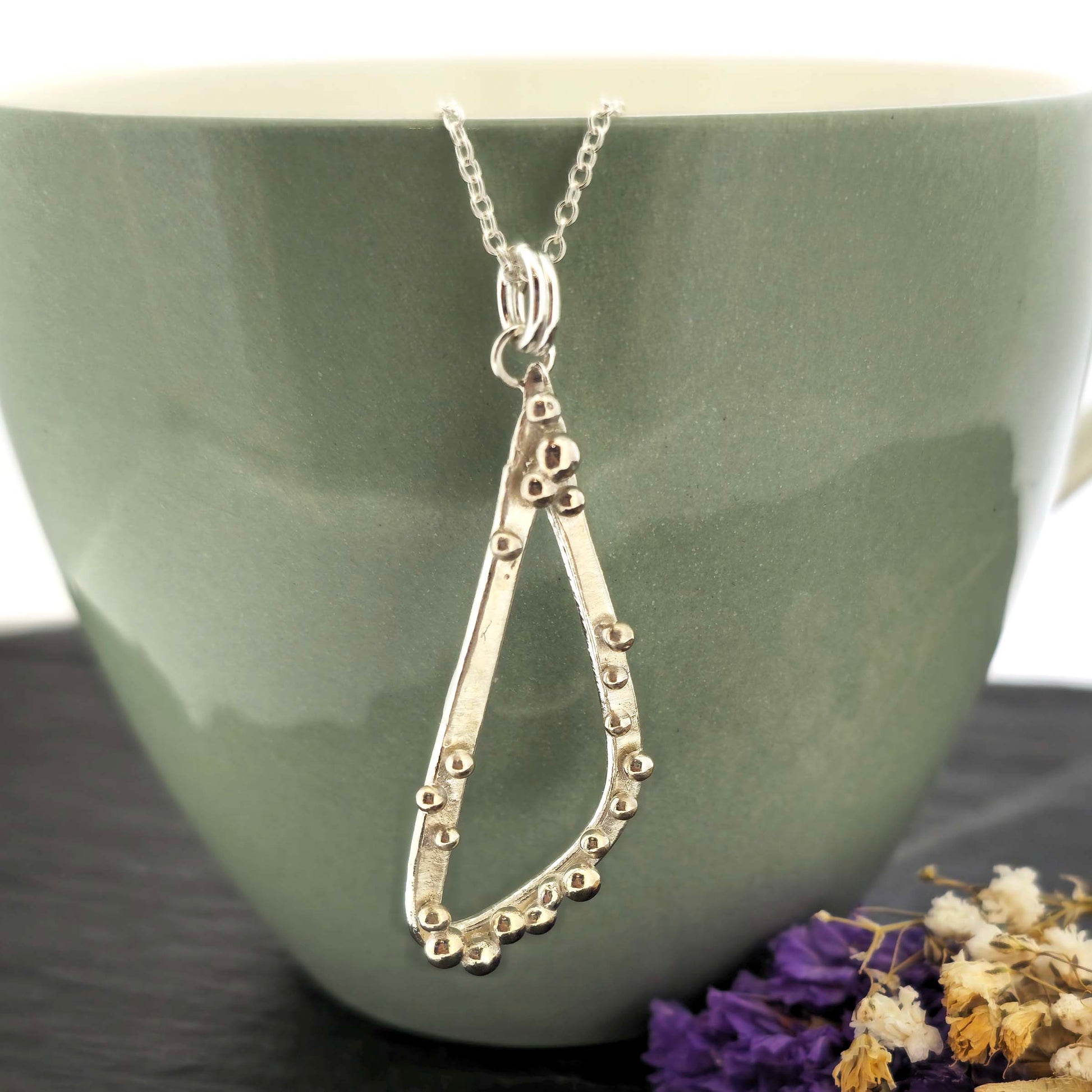 A silver asymmetrical pendant with a soft open triangle shape and silver balls, suspended from a silver chain. Shown on a green bowl with dried flowers. 
