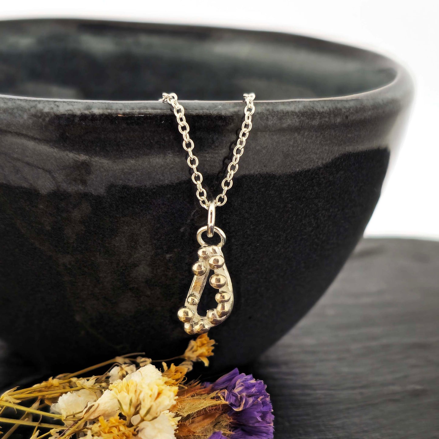 Silver asymmetric open triangle pendant with silver balls on a silver chain. Shown on a black bowl with flowers.