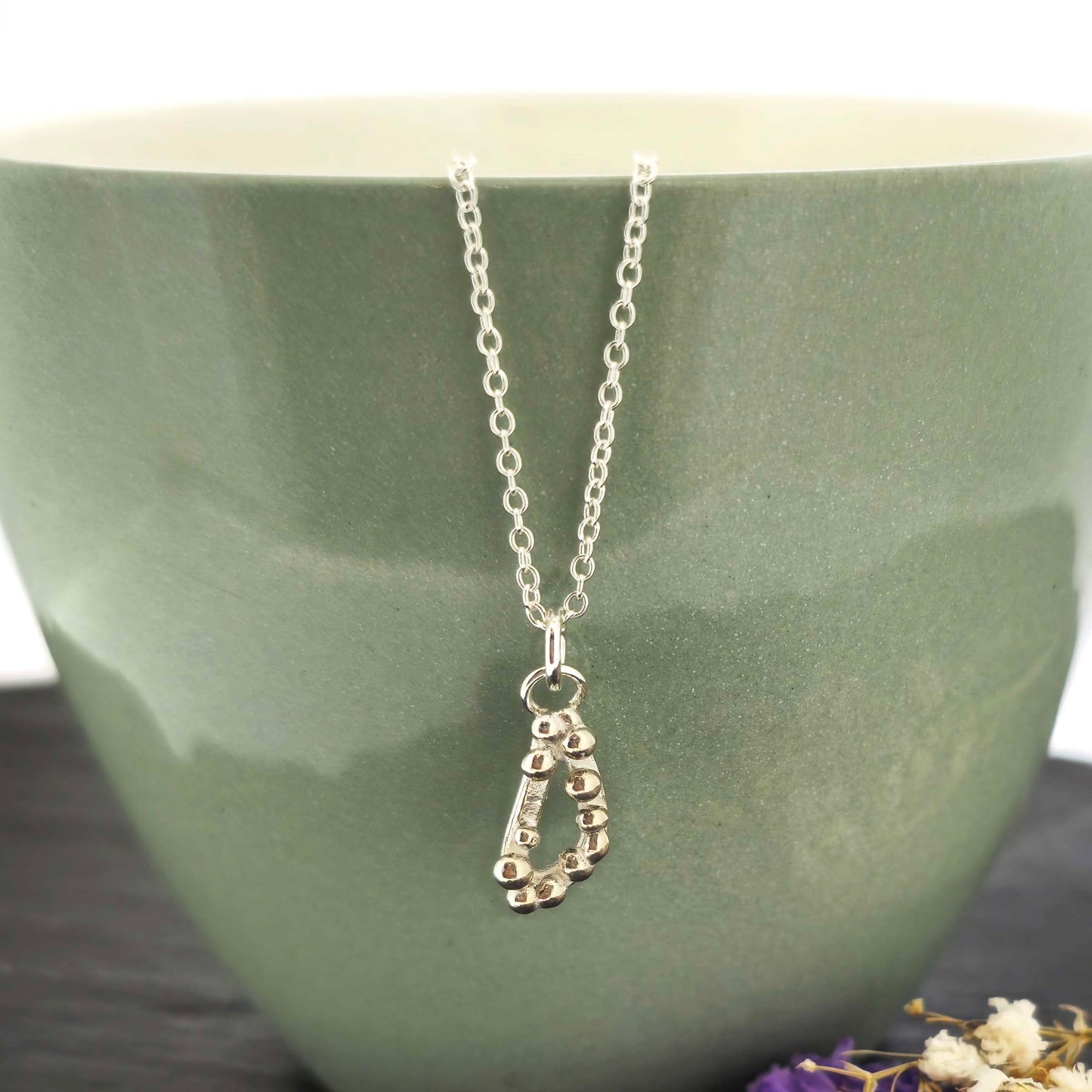 Silver asymmetric open triangle pendant with silver balls on a silver chain. Shown on a green bowl with flowers.