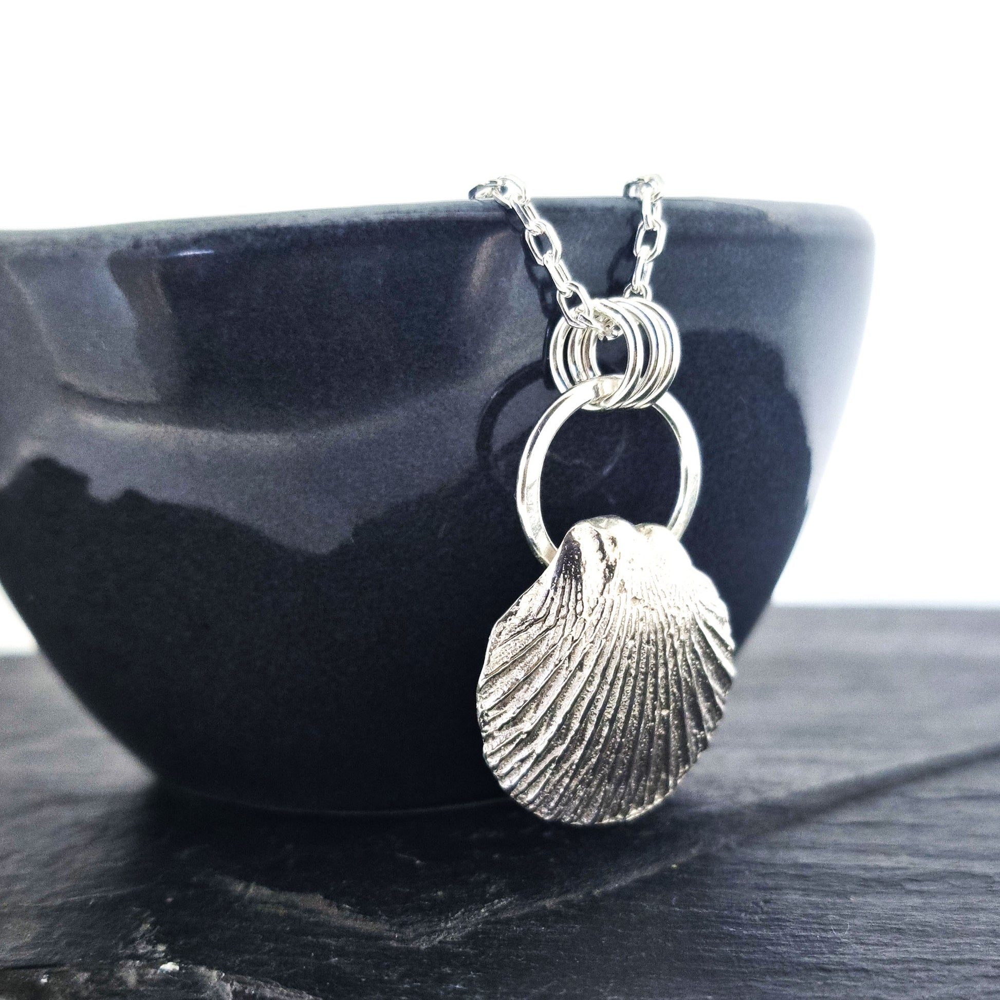 A silver pendant featuring a scallop style shell on a silver circle with 3 loops attaching it to the silver chain. Pictured sideways on a black bowl.