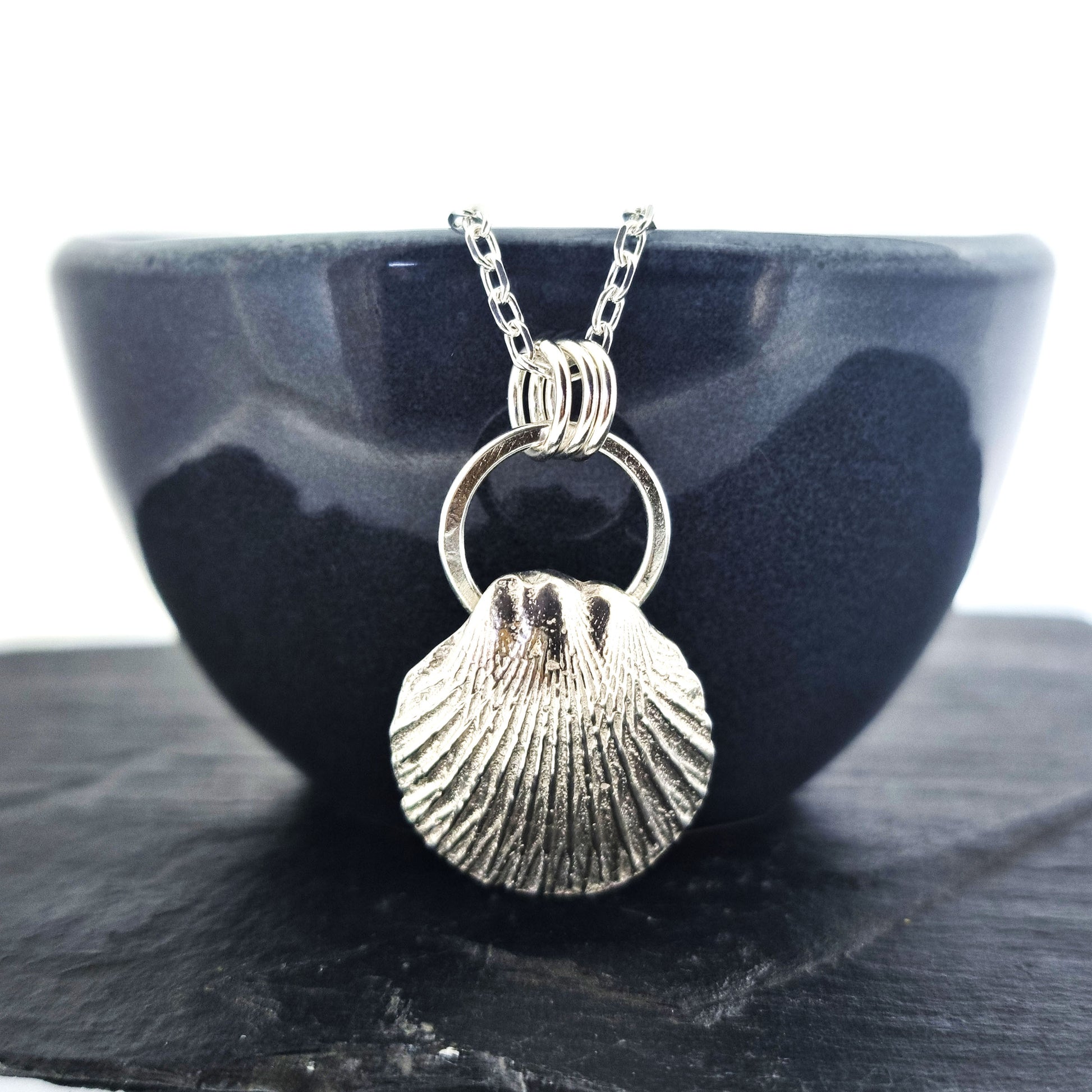 A silver pendant featuring a scallop style shell on a silver circle with 3 loops attaching it to the silver chain. Pictured on a black bowl.