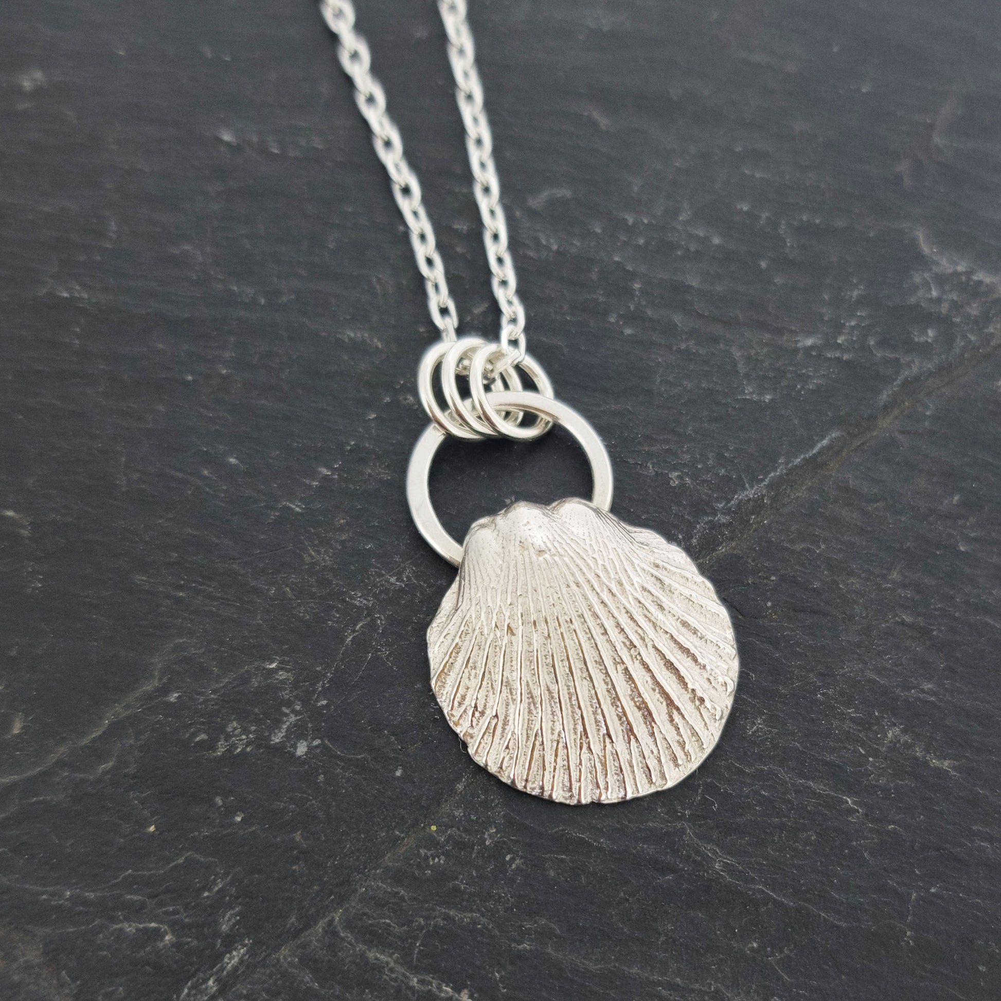 A silver pendant featuring a scallop style shell on a silver circle with 3 loops attaching it to the silver chain. Pictured on slate.