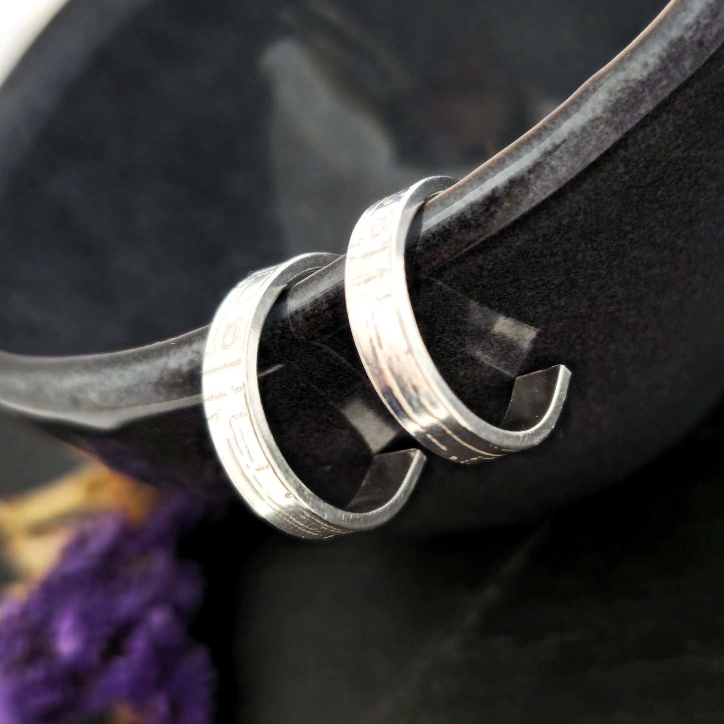 A pair of silver flat hoop earrings with a square and line pattern on. Pictured on the edge of a black bowl.