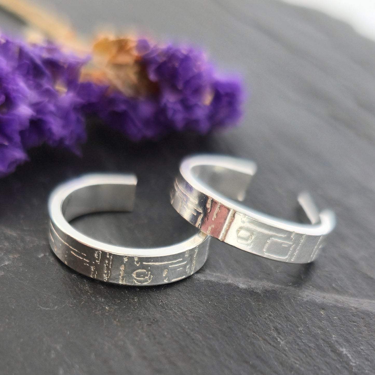 A pair of silver flat hoop earrings with a square and line pattern on. Pictured on slate with purple flowers.