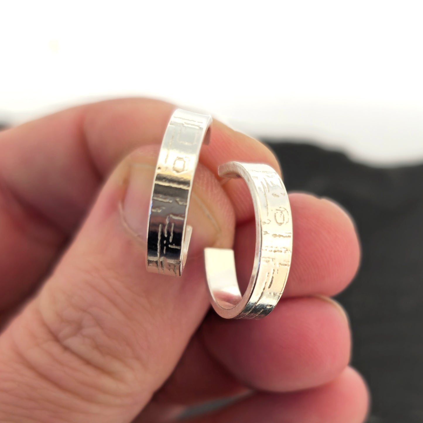 A pair of silver flat hoop earrings with a square and line pattern on. Held between fingers against a blurred background.