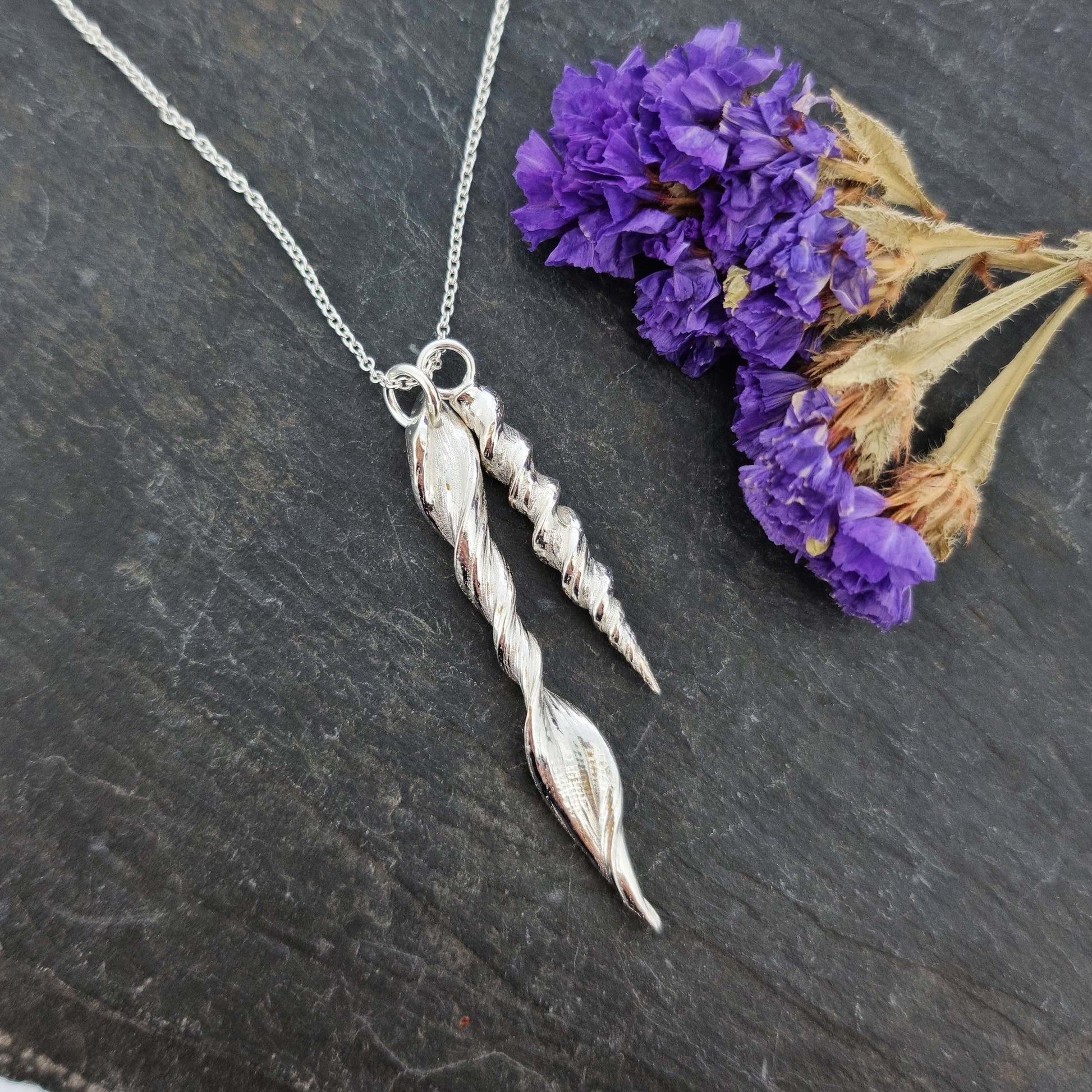 Two silver twist pendants of different sizes suspended from a silver chain. Pictured with flowers.