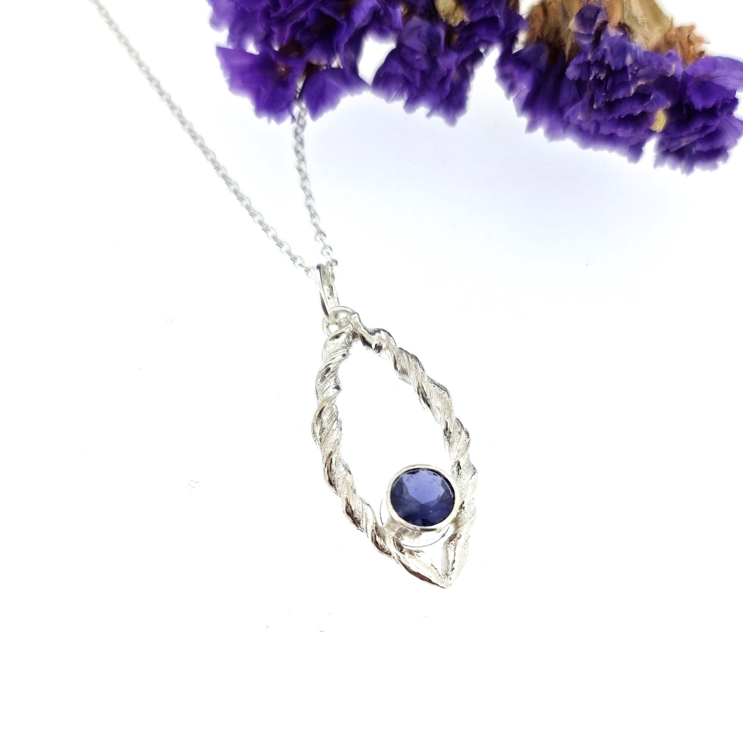 Silver pendant featuring 2 twists of silver making an open boat shape with a purple blue iolite gemstone set in the bottom point. On a silver chain. Pictured with flowers.