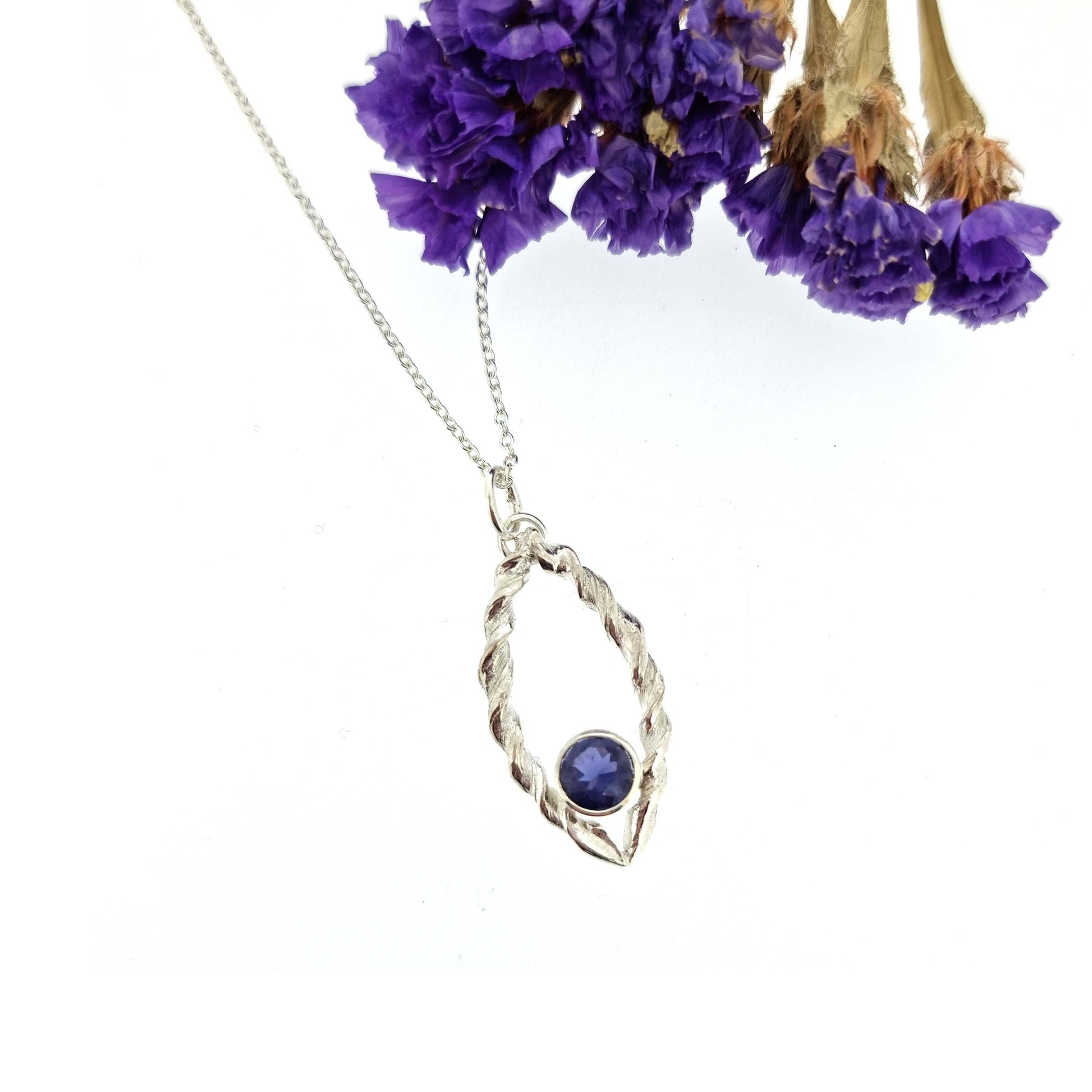 Silver pendant featuring 2 twists of silver making an open boat shape with a purple blue iolite gemstone set in the bottom point. On a silver chain. Pictured with flowers.