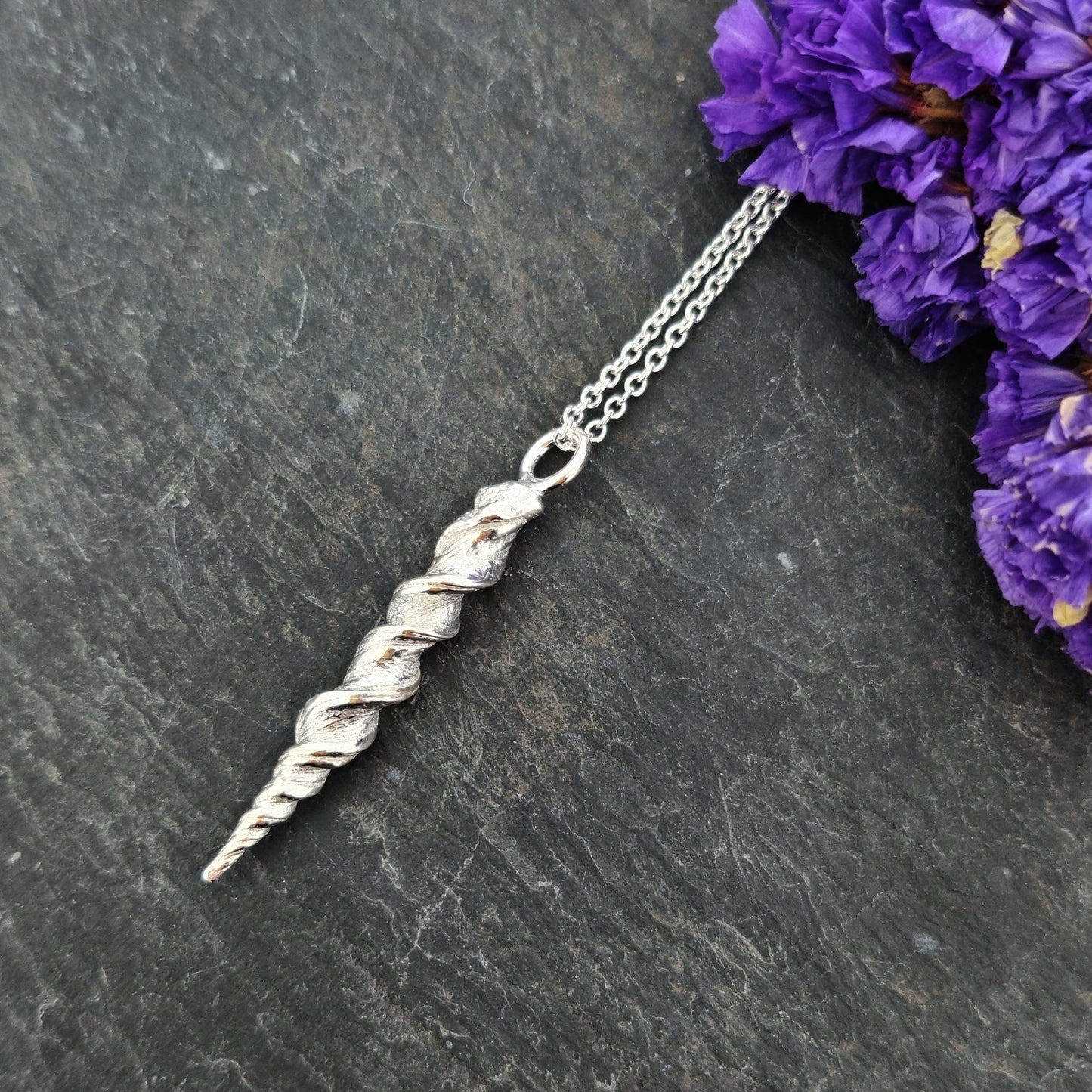 A silver twist pendant with rustic detail on a silver chain. Pictured with flowers.