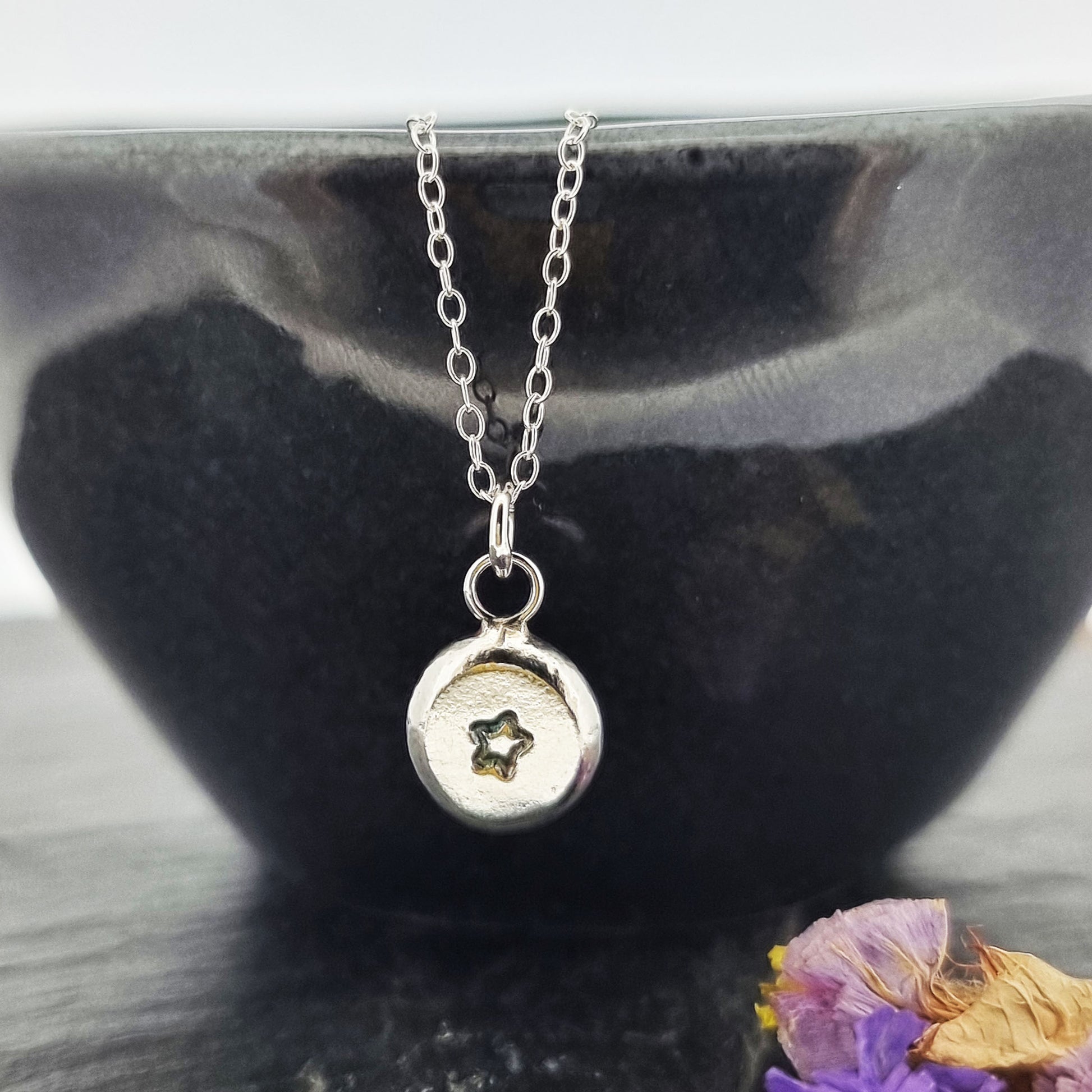 A silver flat pebble pendant with a black 5 pointed star on it. On a silver chain. Pictured on a bowl.