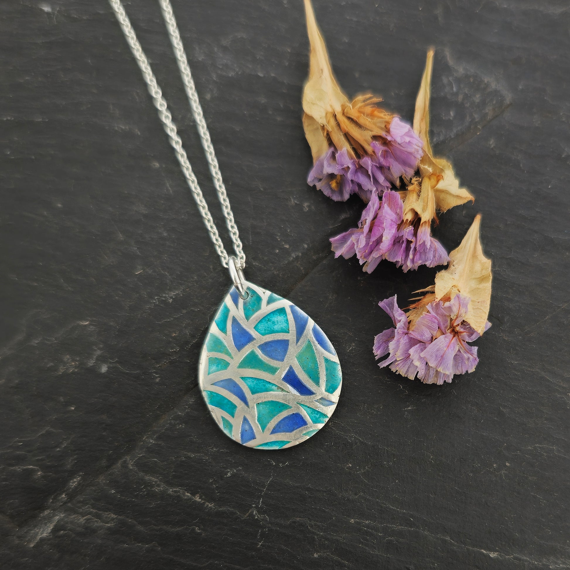 A teardrop shaped silver pendant with geometric cells of blue, green and turquoise enamel. On a silver chain. Pictured on slate with flowers.