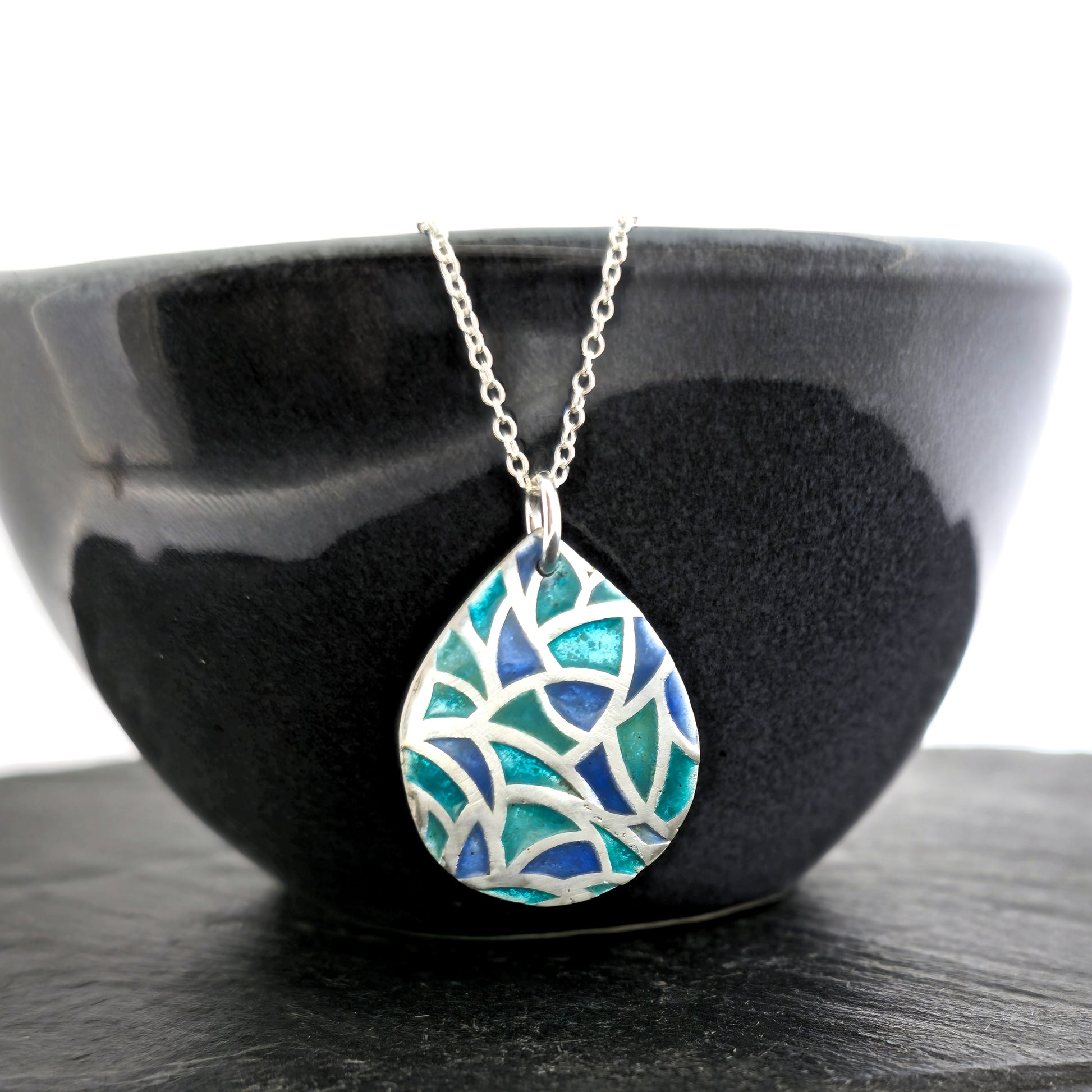 A teardrop shaped silver pendant with geometric cells of blue, green and turquoise enamel. On a silver chain. Pictured on a black bowl.