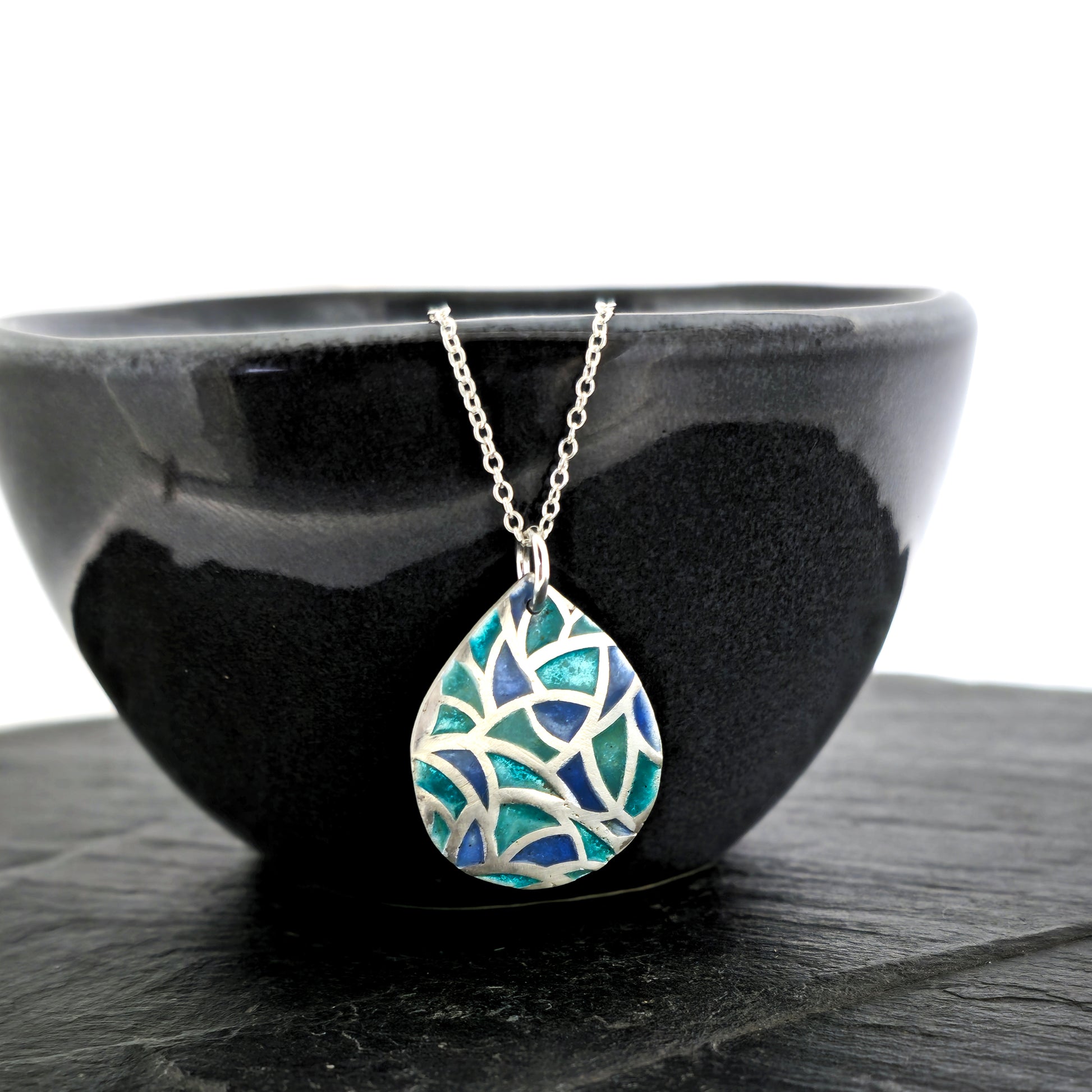 A teardrop shaped silver pendant with geometric cells of blue, green and turquoise enamel. On a silver chain. Pictured on a black bowl.