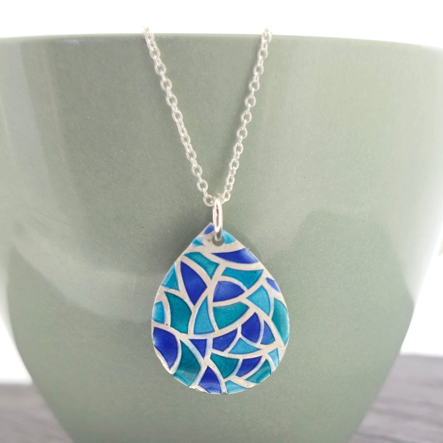 A teardrop shaped silver pendant with geometric cells of blue, green and turquoise enamel. On a silver chain. Pictured on a green bowl.