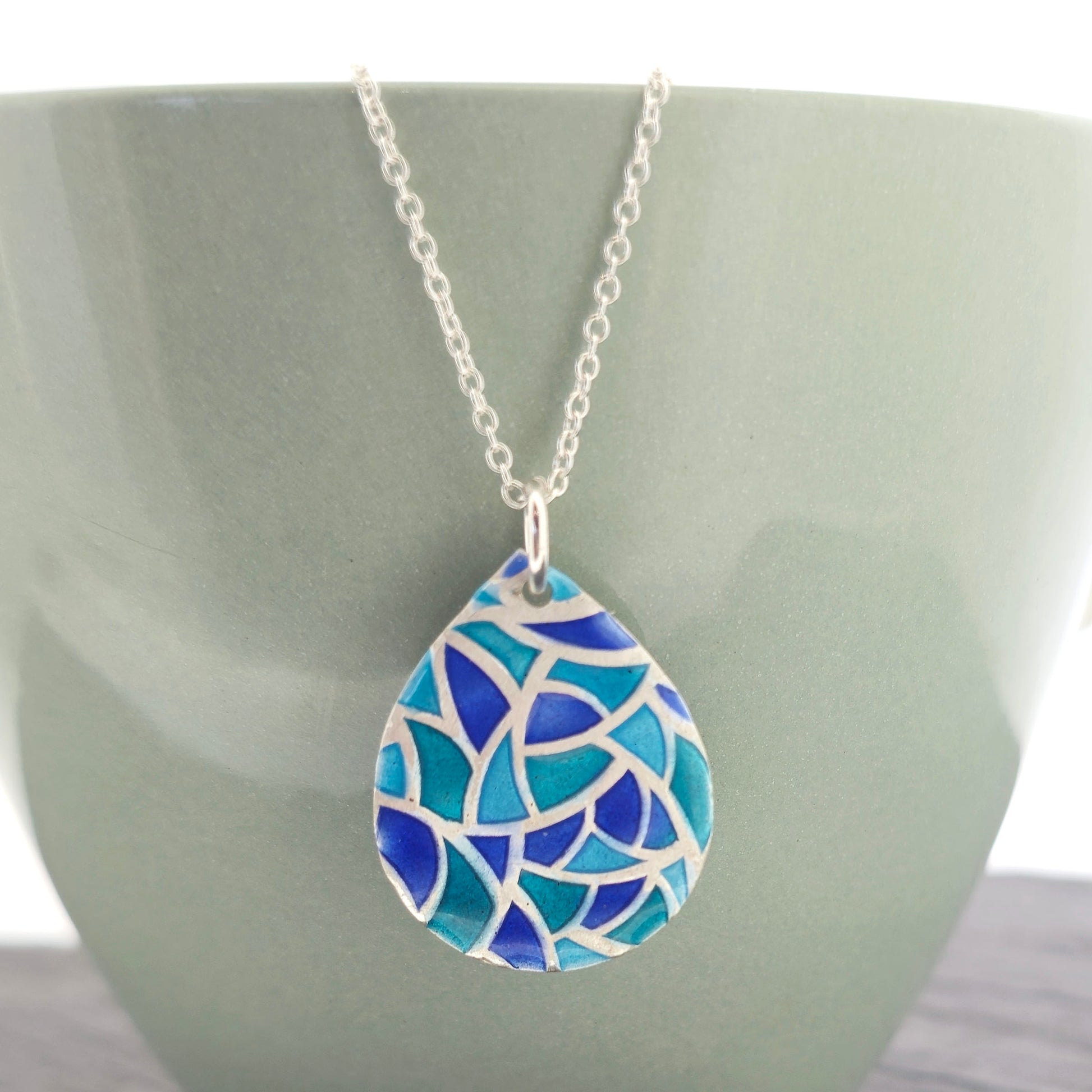 A teardrop shaped silver pendant with geometric cells of blue, green and turquoise enamel. On a silver chain. Pictured on a green bowl.