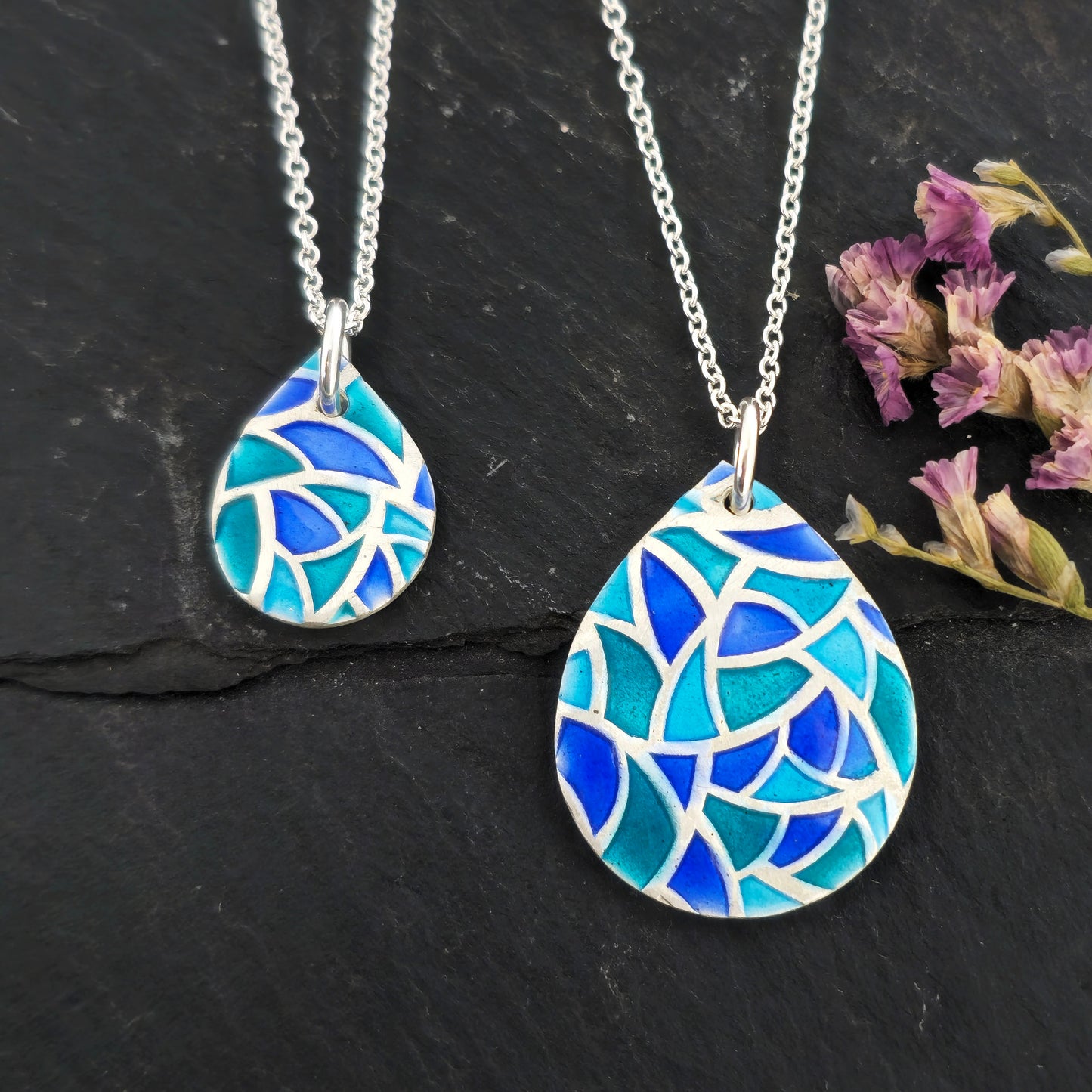 Two teardrop shaped silver pendants with geometric cells of blue, green and turquoise enamel. On a silver chain. Pictured on a black surface with dried flowers.