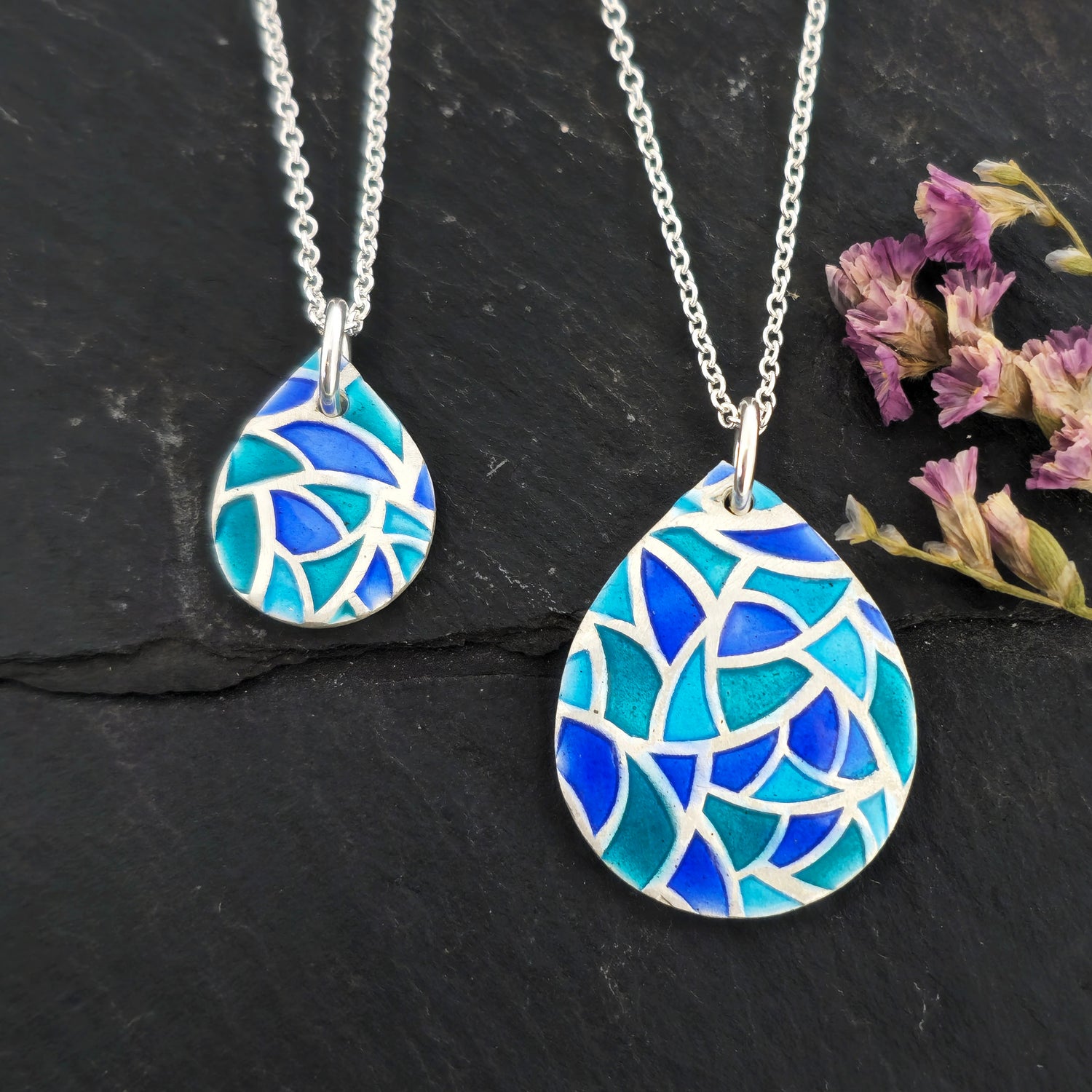 Two teardrop shaped silver pendants with geometric cells of blue, green and turquoise enamel. On a silver chain. Pictured on a black surface with dried flowers.