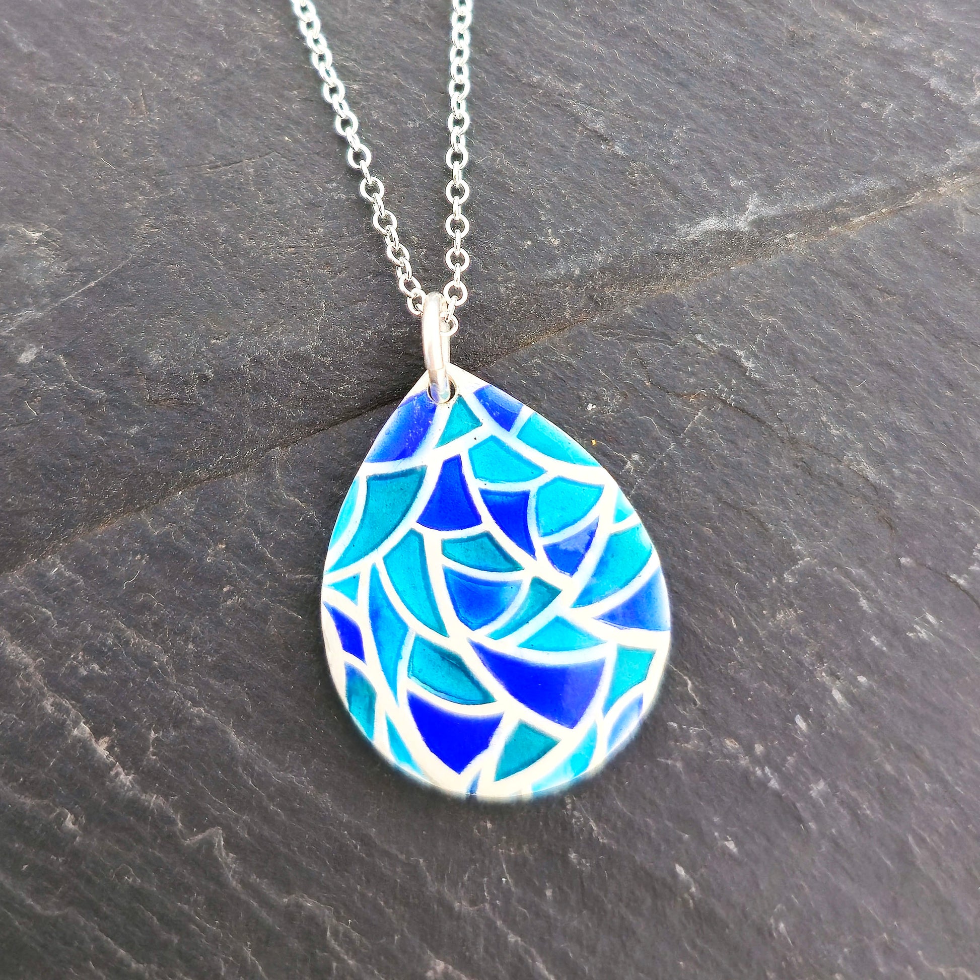 A teardrop shaped silver pendant with geometric cells of blue, green and turquoise enamel. On a silver chain. Pictured on a black background. Handmade. Mijoux Jewellery.