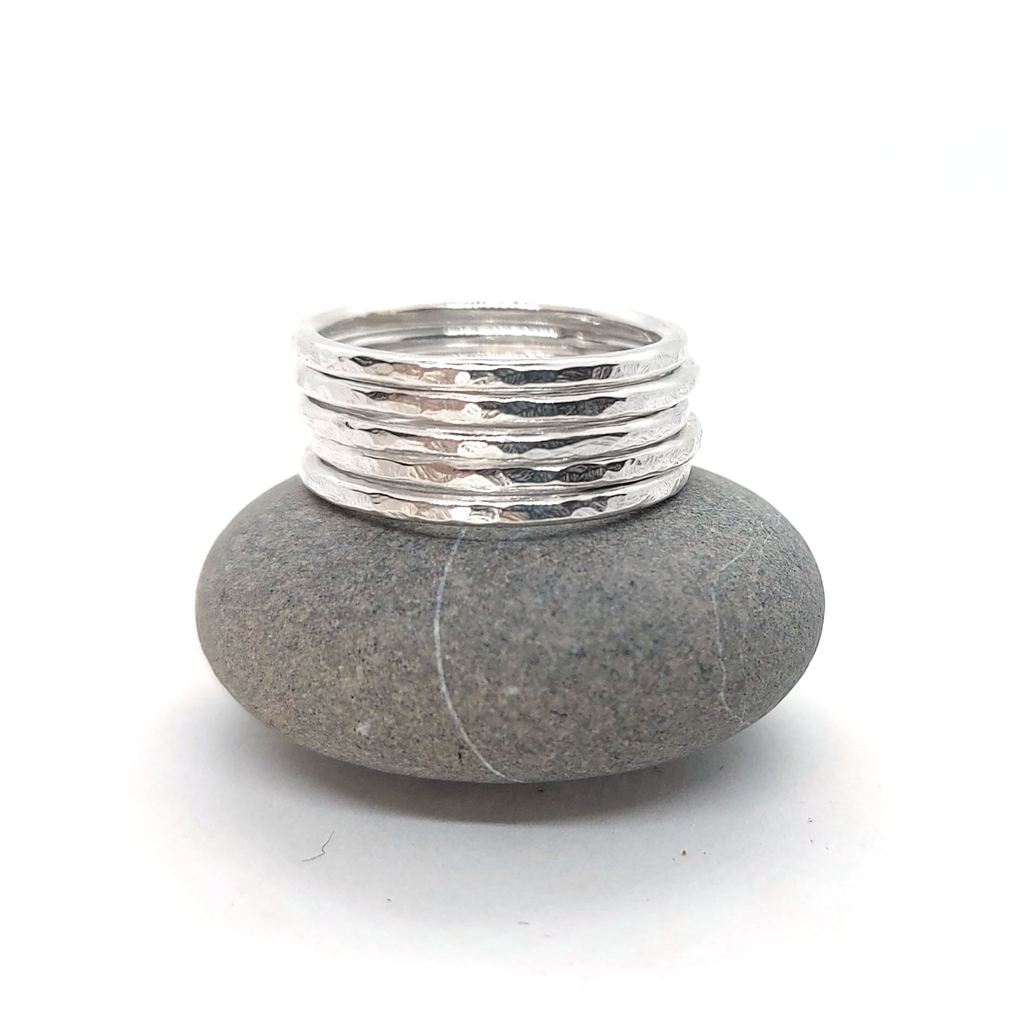 A set of 5 thin silver stacking rings with a hammered finish. Shown pictured on a pebble.
