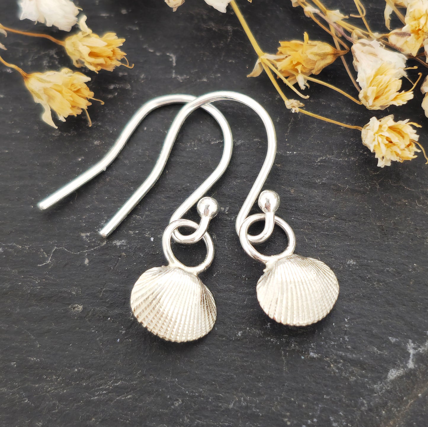 Silver cockle shell drop earrings on black background with flowers. Handmade. Mijoux Jewellery.