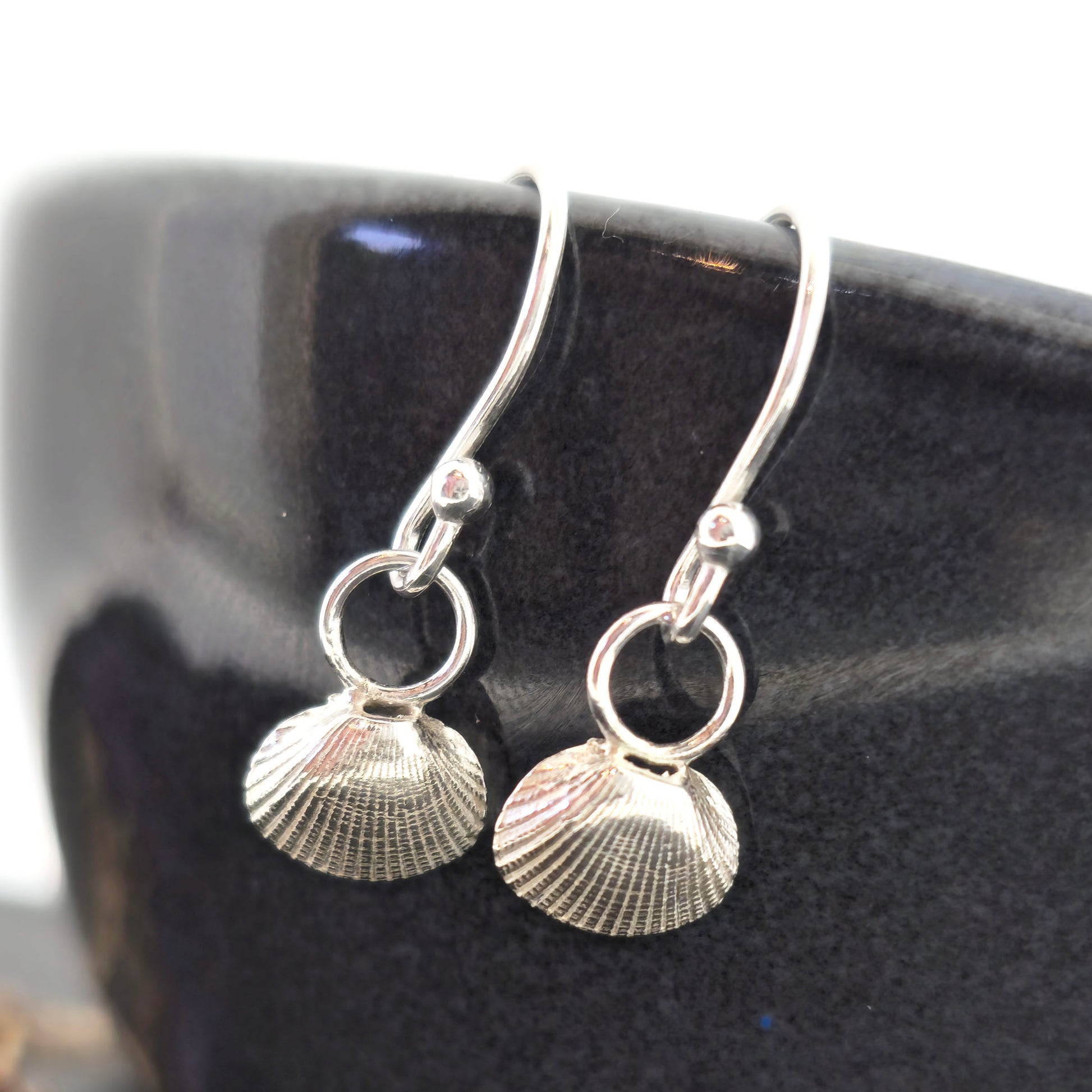 Silver cockle shell drop earrings on black bowl. Handmade. Mijoux Jewellery.