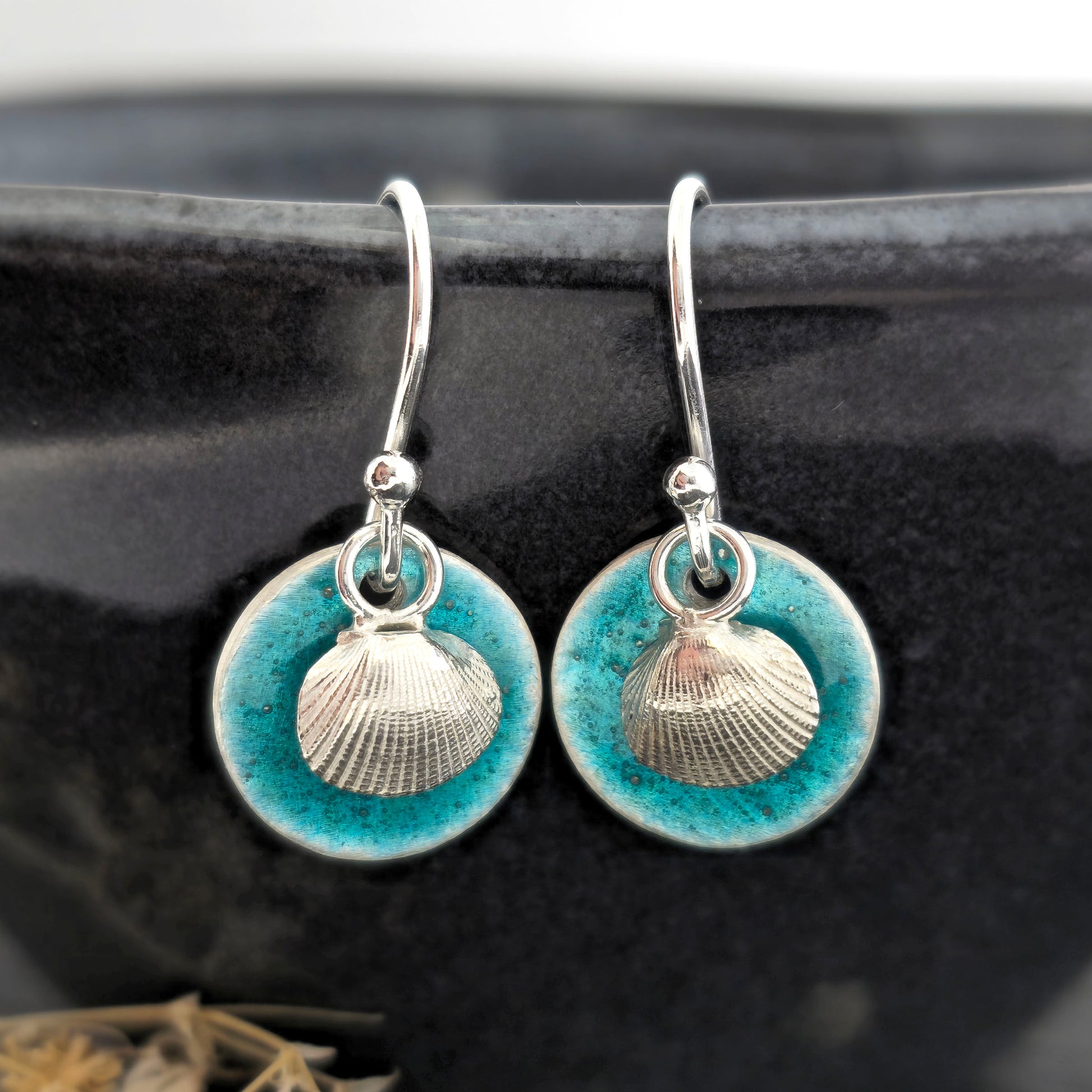 A pair of handmade silver earrings with a turquoise enamel disc and a silver cockle shell on front. On a black background. Mijoux Jewellery.