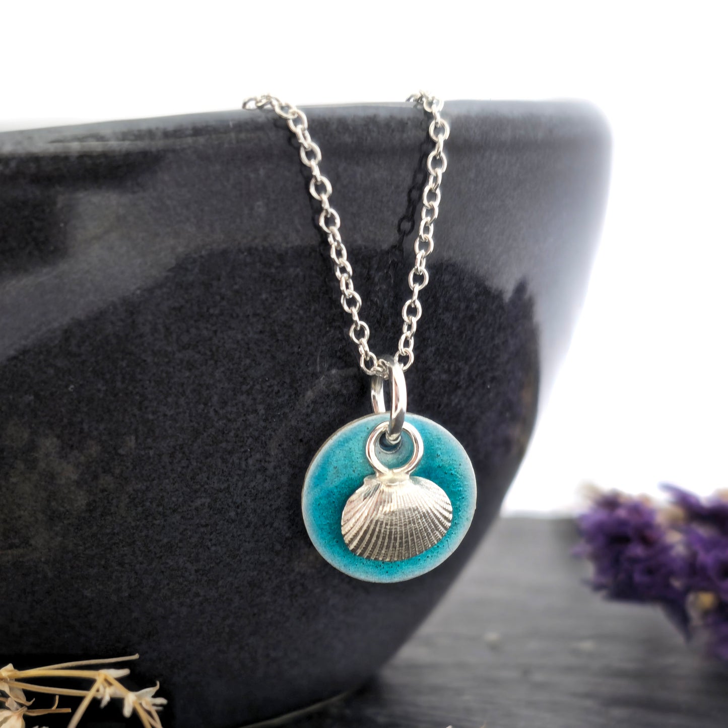 A silver pendant with a turquoise enamel disc and a silver cockle shell in front. On a silver chain. Shown on a black bowl with flowers. Handmade. Mijoux Jewellery.