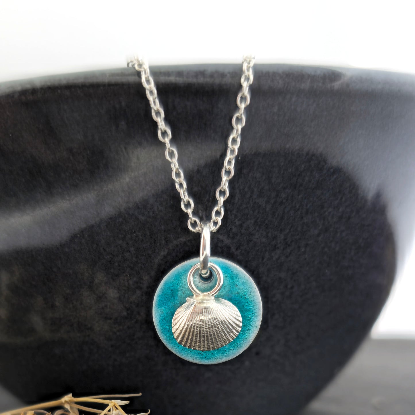 A silver pendant with a turquoise enamel disc and a silver cockle shell in front. On a silver chain. Shown on a black bowl. Handmade. Mijoux Jewellery.