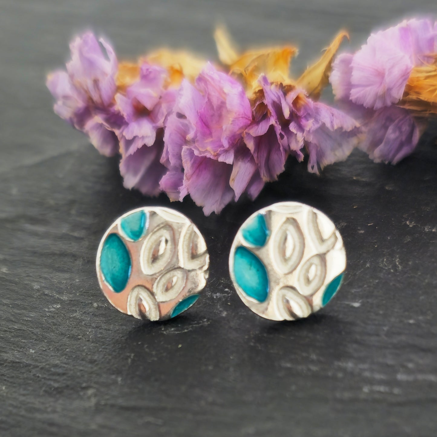 Round silver stud earrings with a leaf shape pattern. Some leaves are filled with turquoise enamel. Pictured on slate with flowers.