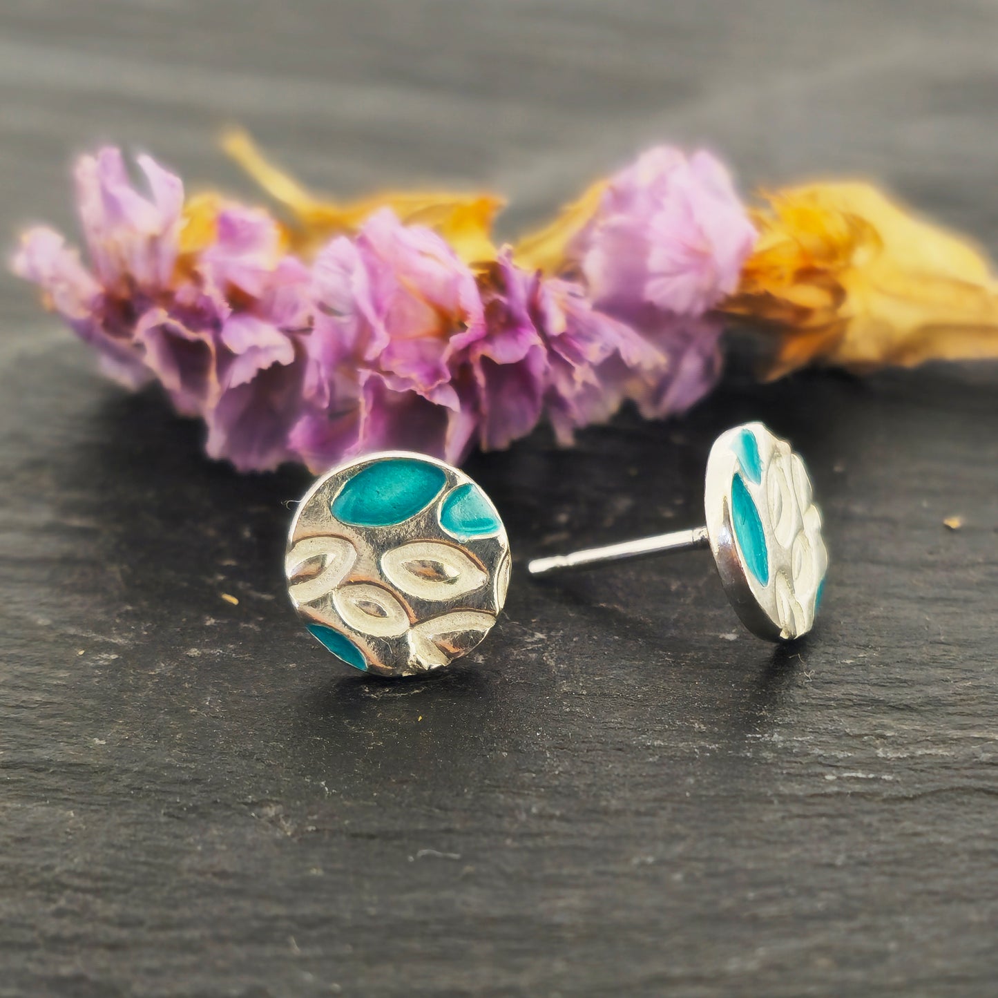 Round silver stud earrings with a leaf shape pattern. Some leaves are filled with turquoise enamel. Pictured on slate with flowers.