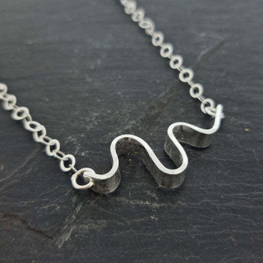 A silver necklace with an open chain and a wave of silver at the center. Shown on slate.