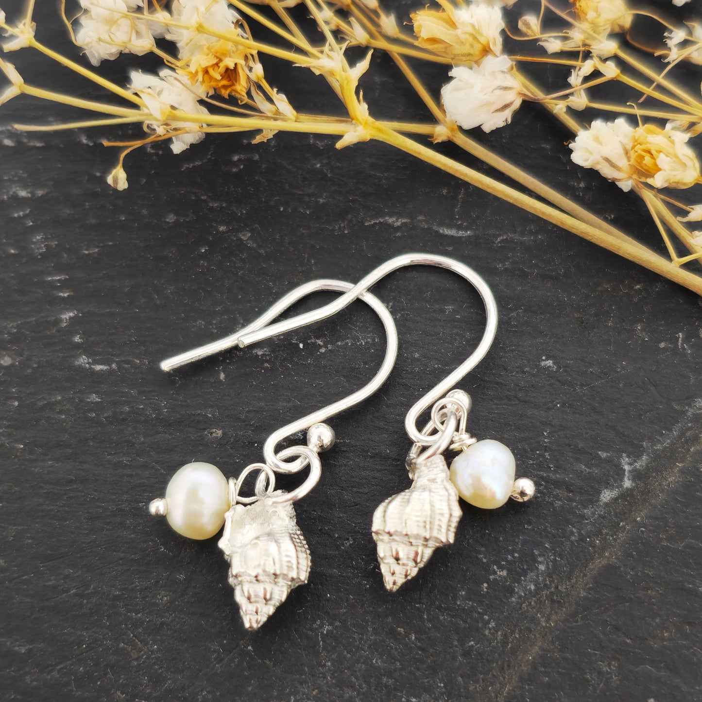 A pair of silver periwinkle shell and freshwater pearl drop earrings on a black background with flowers. Handmade. Mijoux Jewellery.