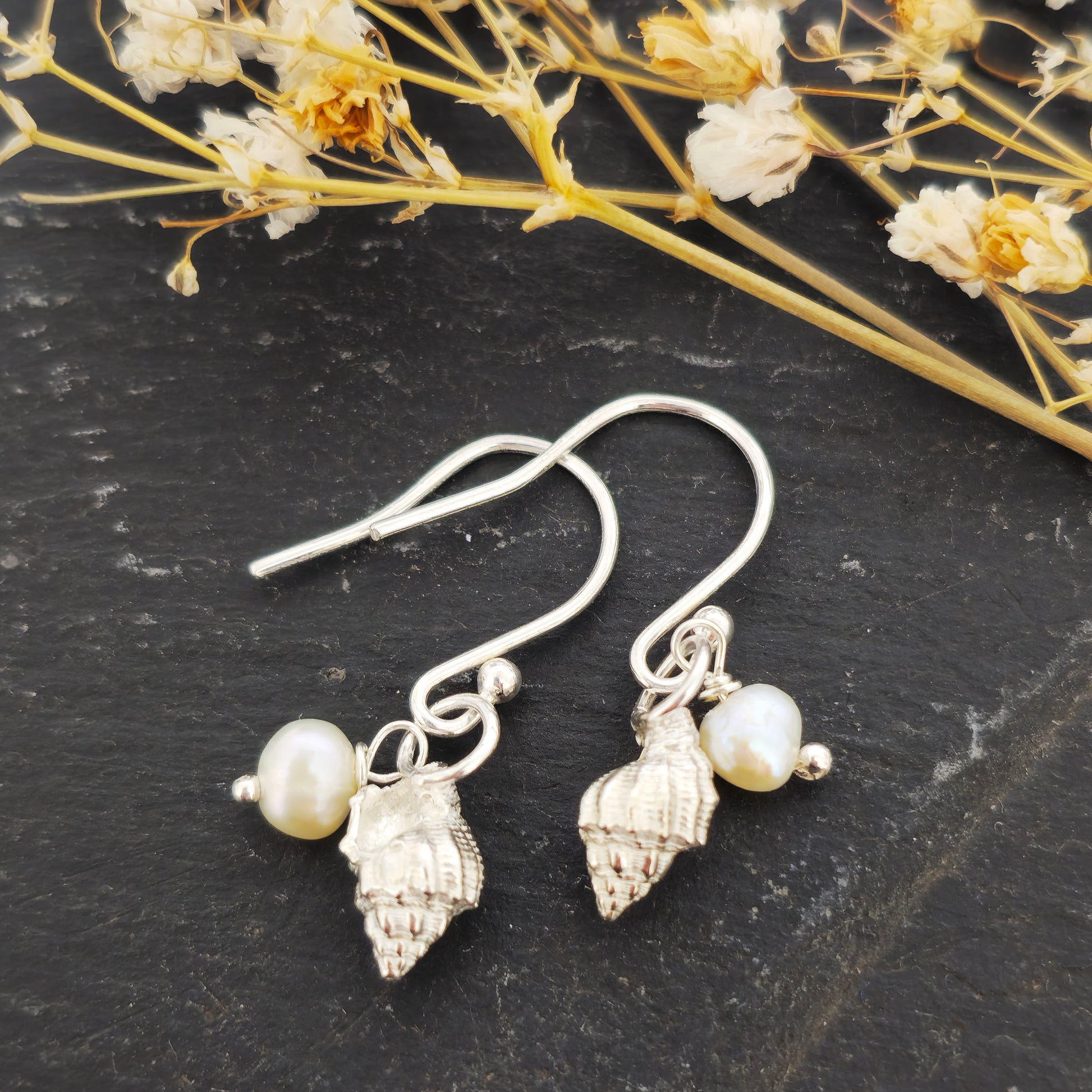 A pair of silver periwinkle shell and freshwater pearl drop earrings on a black background with flowers. Handmade. Mijoux Jewellery.
