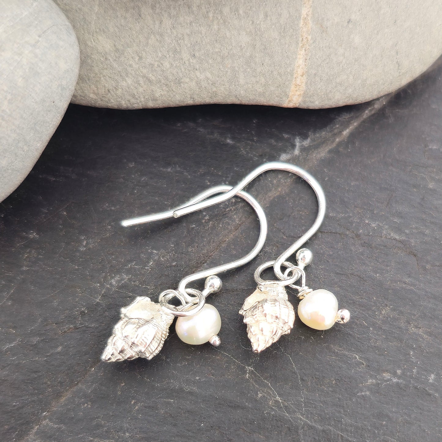 A pair of silver periwinkle shell and freshwater pearl drop earrings on a black background with pebbles. Handmade. Mijoux Jewellery.