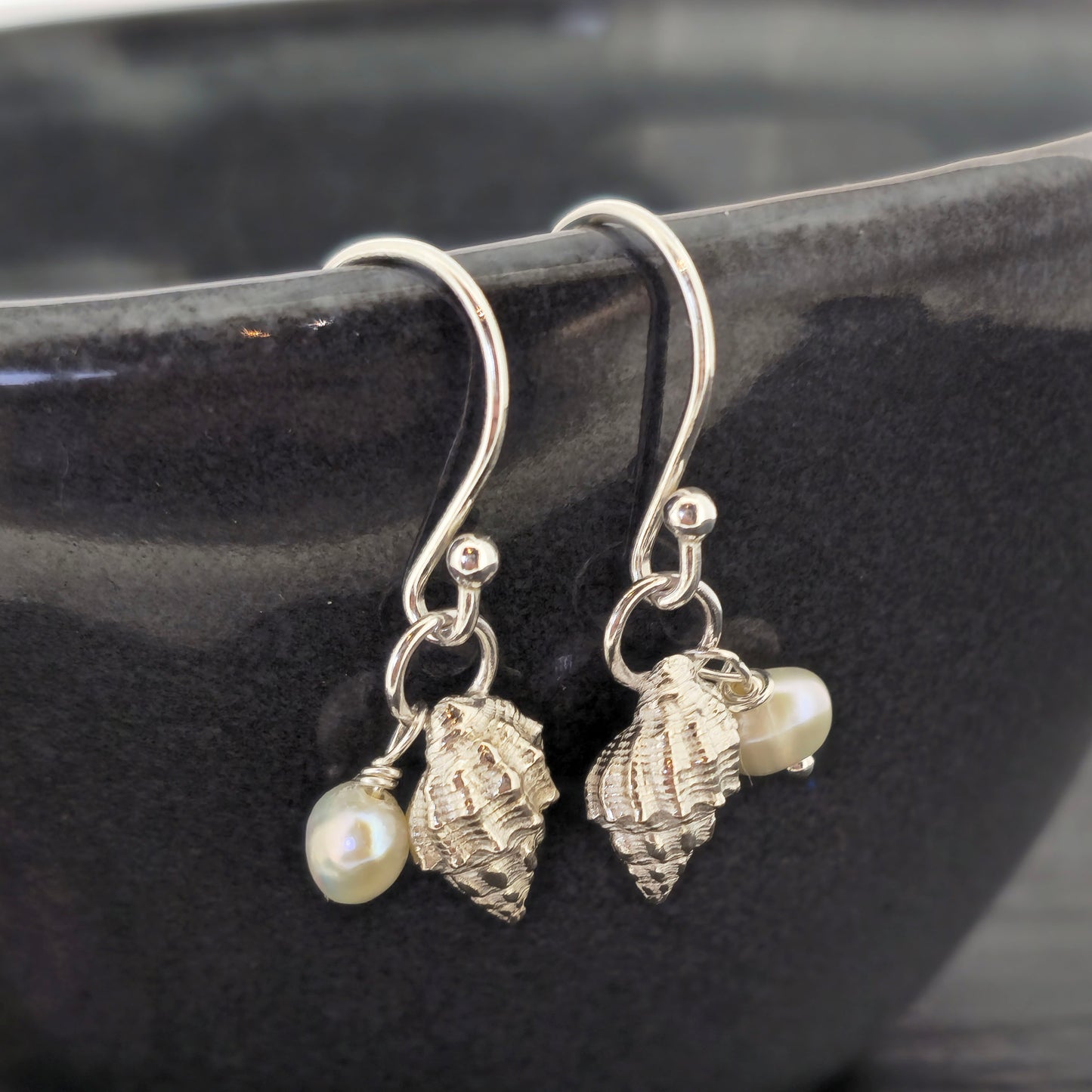 A pair of silver periwinkle shell and freshwater pearl drop earrings on a black bowl. Handmade. Mijoux Jewellery.