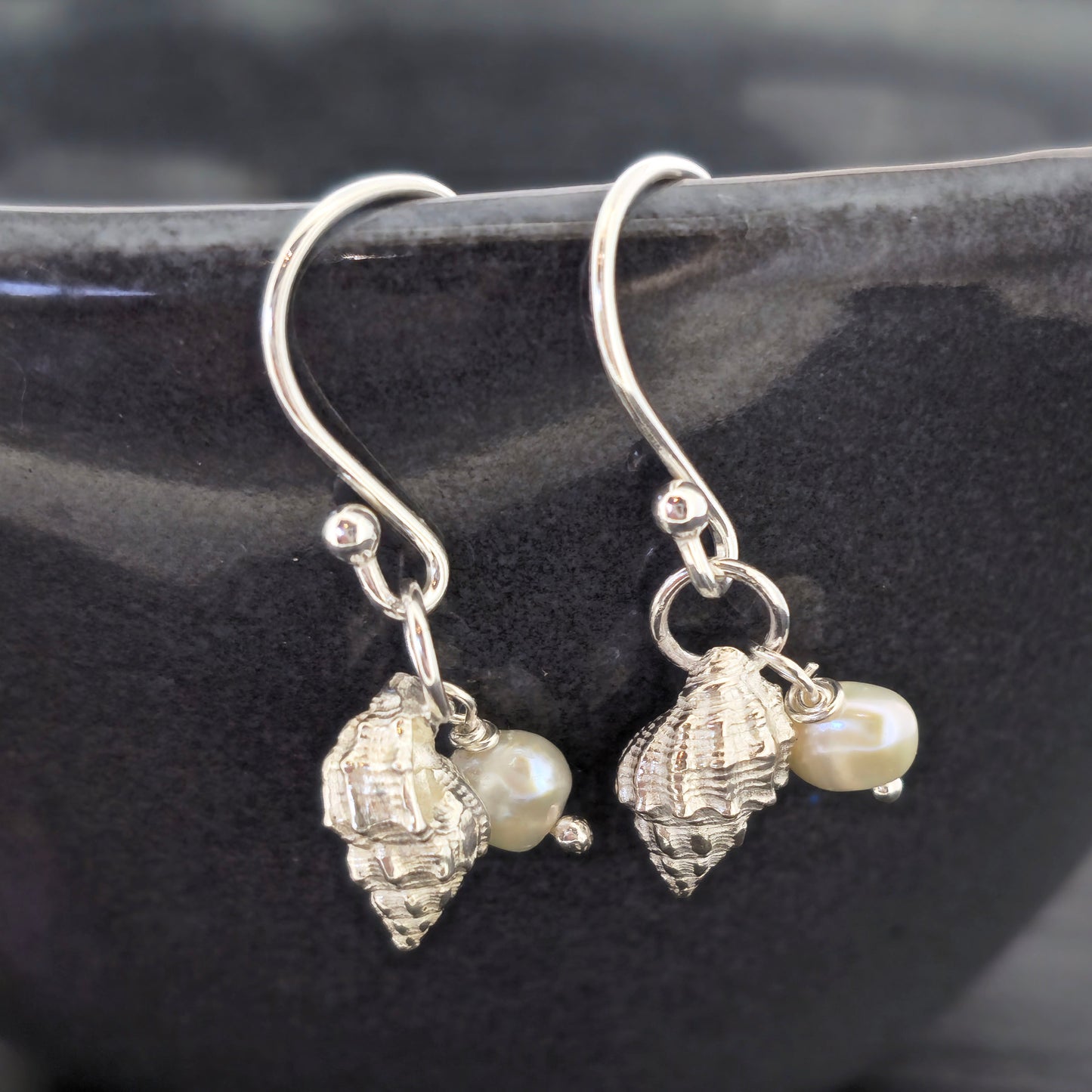 A pair of silver periwinkle shell and freshwater pearl drop earrings on a black bowl. Handmade. Mijoux Jewellery.