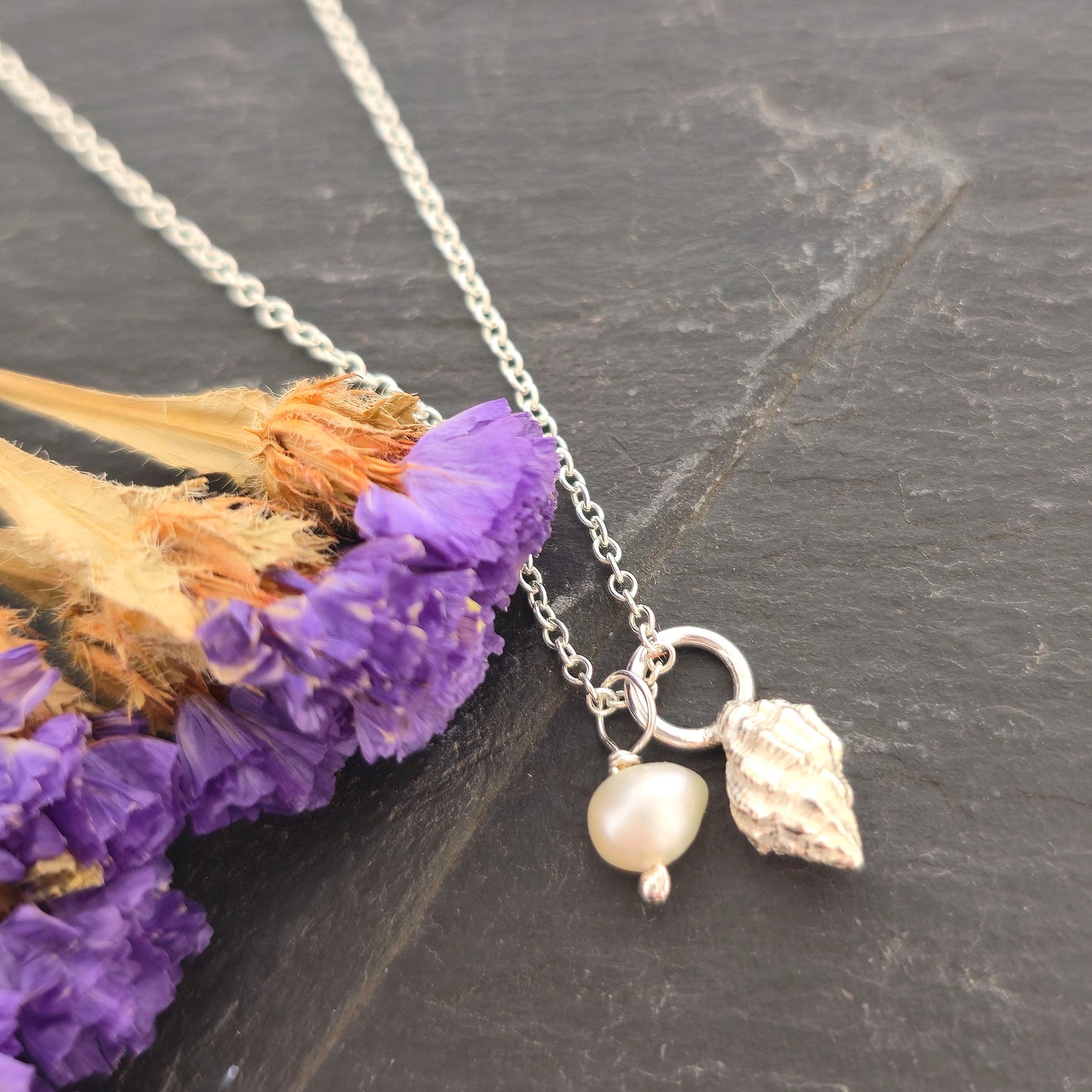 A silver periwinkle shell pendant with a cream coloured pearl suspended from a silver chain. On a black background with purple flowers. Handmade. Mijoux Jewellery.