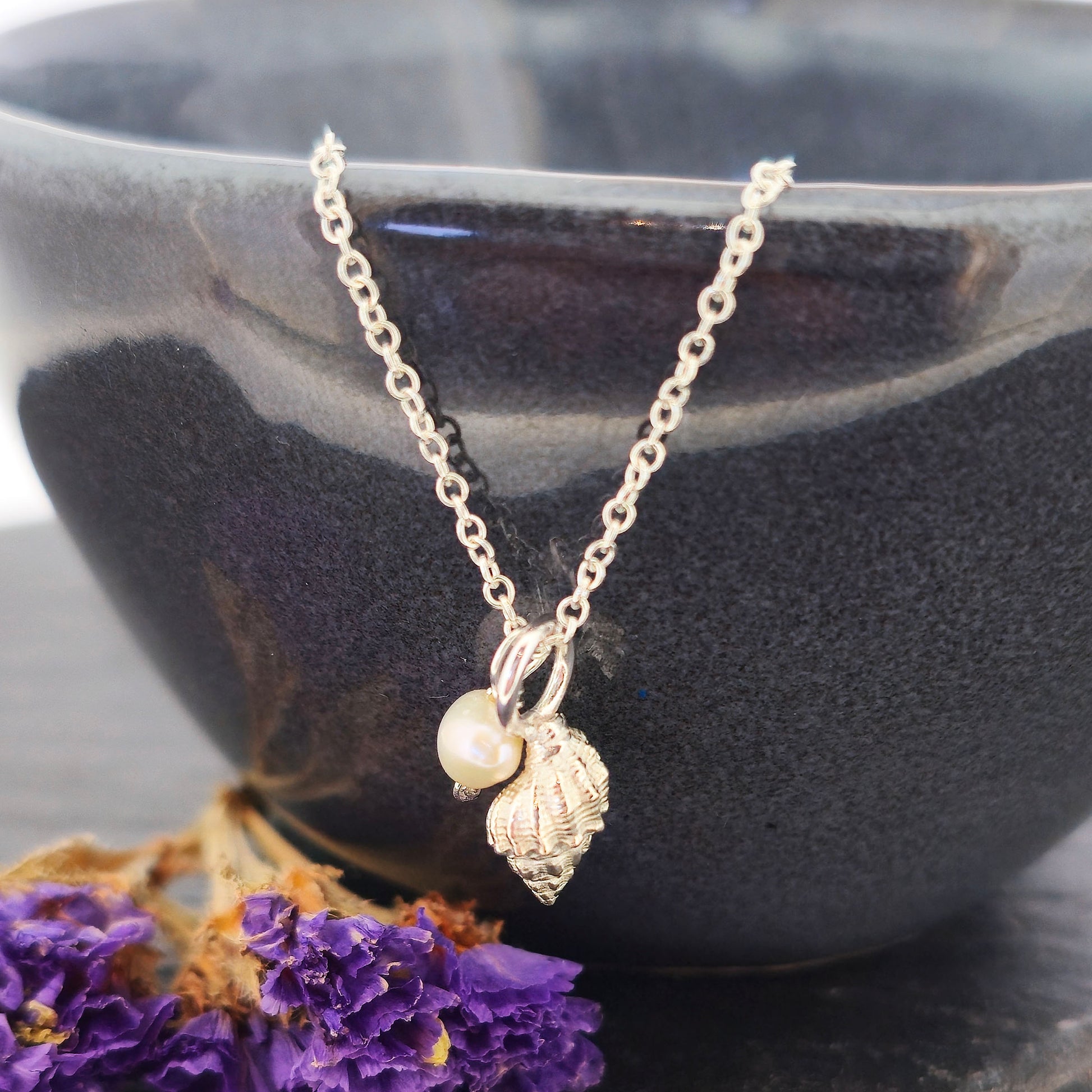 A silver periwinkle shell pendant with a cream coloured pearl suspended from a silver chain. On a black bowl with purple flowers. Handmade. Mijoux Jewellery.